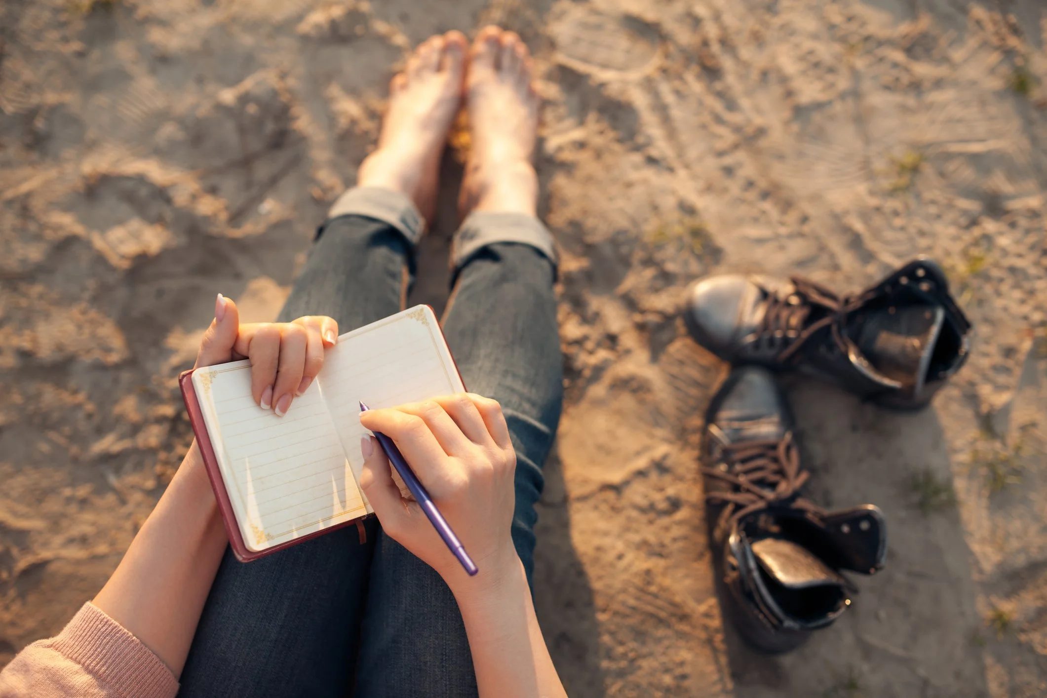 Person sitting on sandy ground at sunset, taking notes in a small notebook, with black boots beside them. Teen or children support, journaling, therapy support.