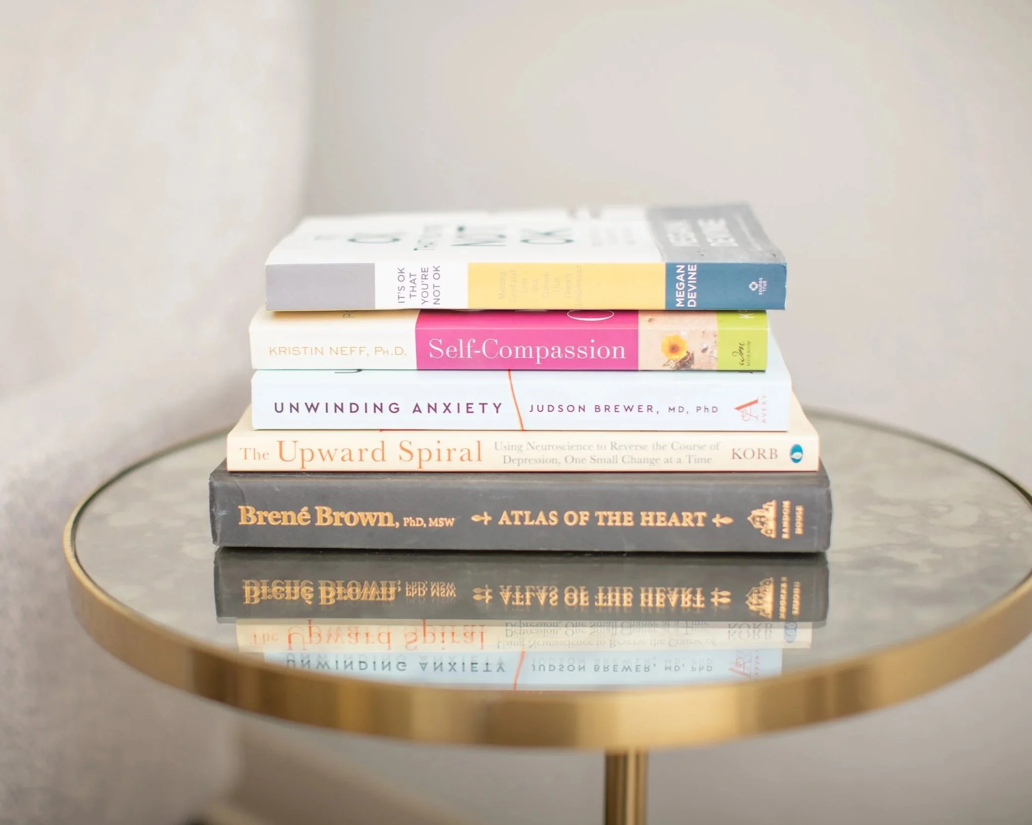 Stack of seven self-help and psychology books on a glass-topped side table with a gold rim, reflected on the surface. Specialties. Resources.