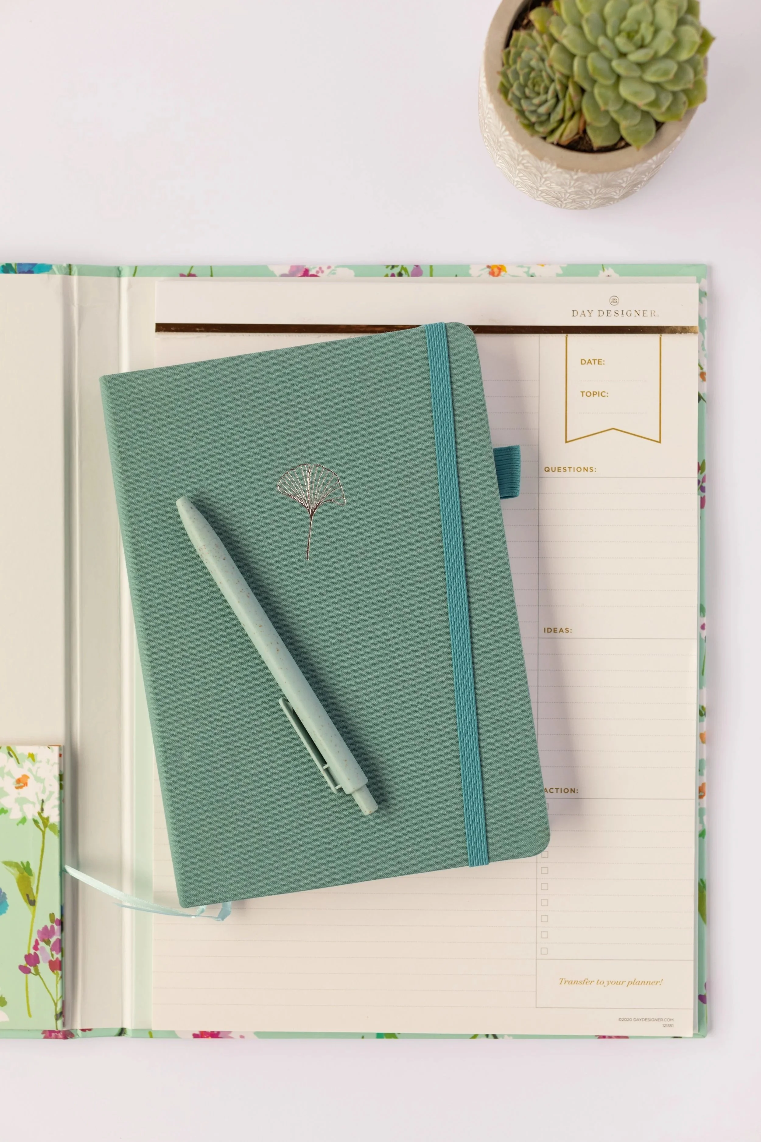 A teal planner with a matching pen resting on top, placed on a floral-patterned notebook and a blank planner page, with a small potted succulent plant nearby on a white surface. Contact Me.