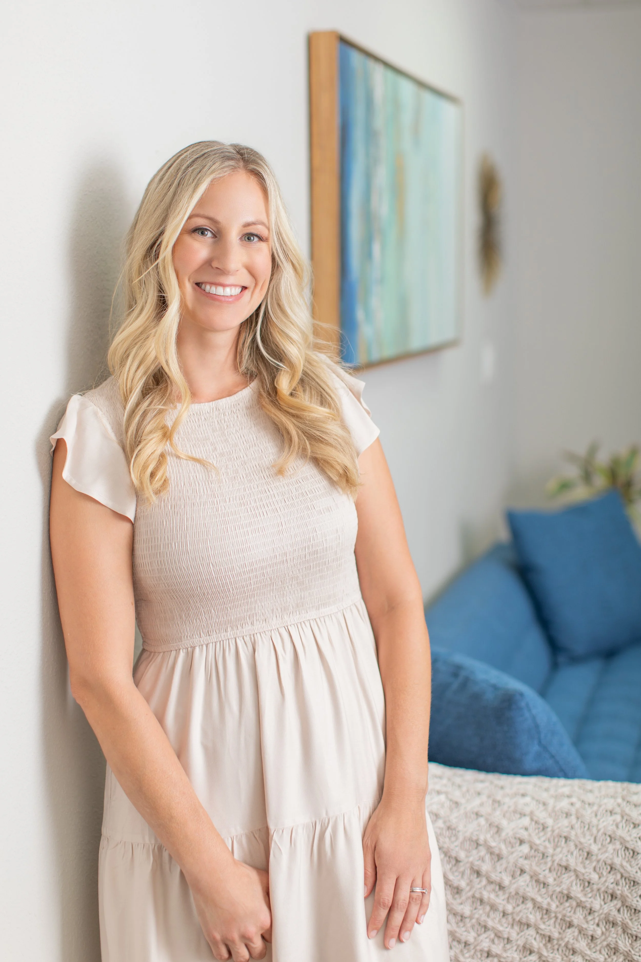A smiling therapist with blonde hair in a light-colored dress, standing and leaning against a white wall in a living room with blue cushions and abstract wall art. Warm and inviting.