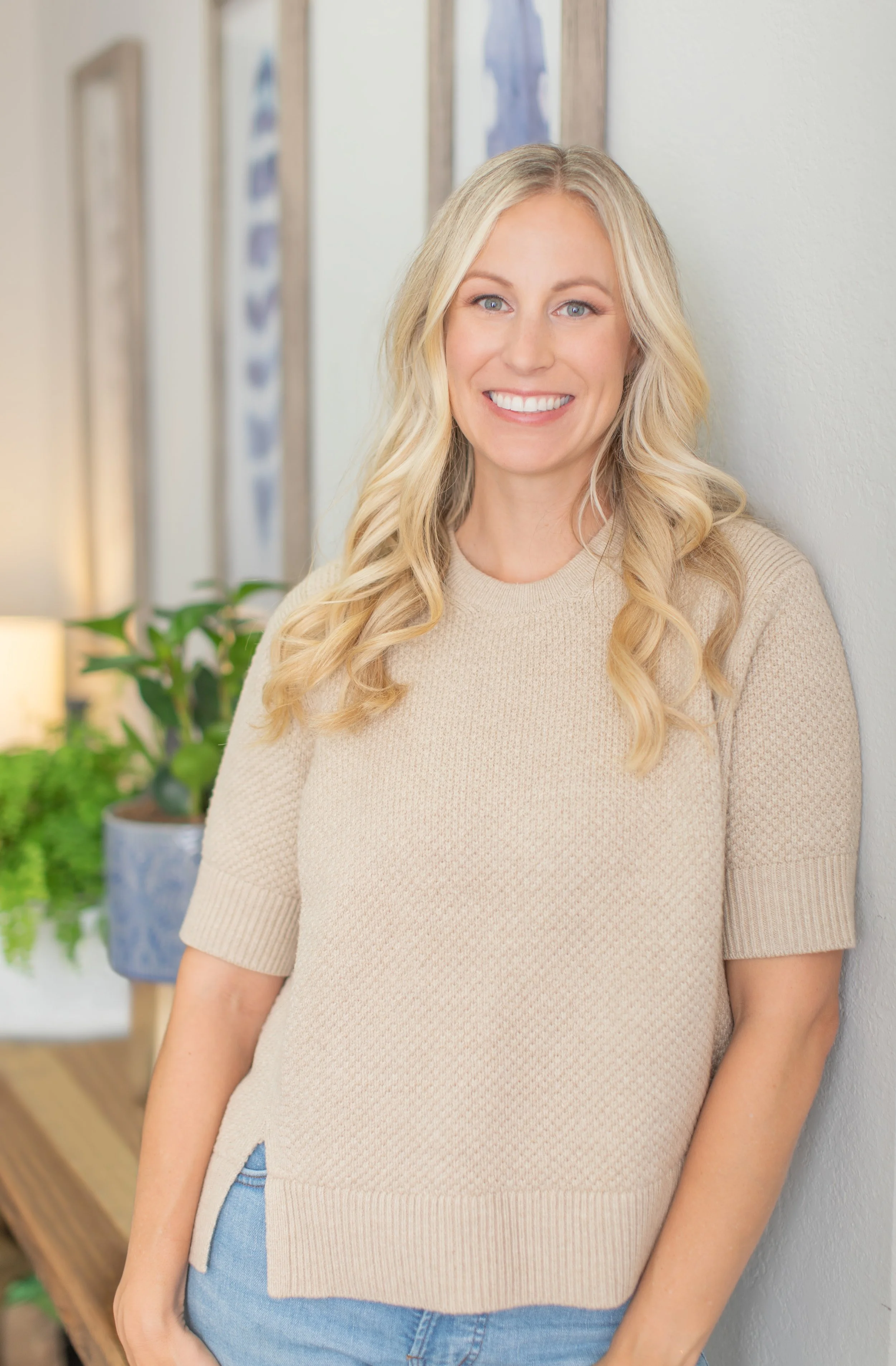 A smiling woman with long blonde hair, wearing a beige knit sweater and jeans, leaning against a wall in a brightly lit indoor setting with plants and framed artwork in the background.