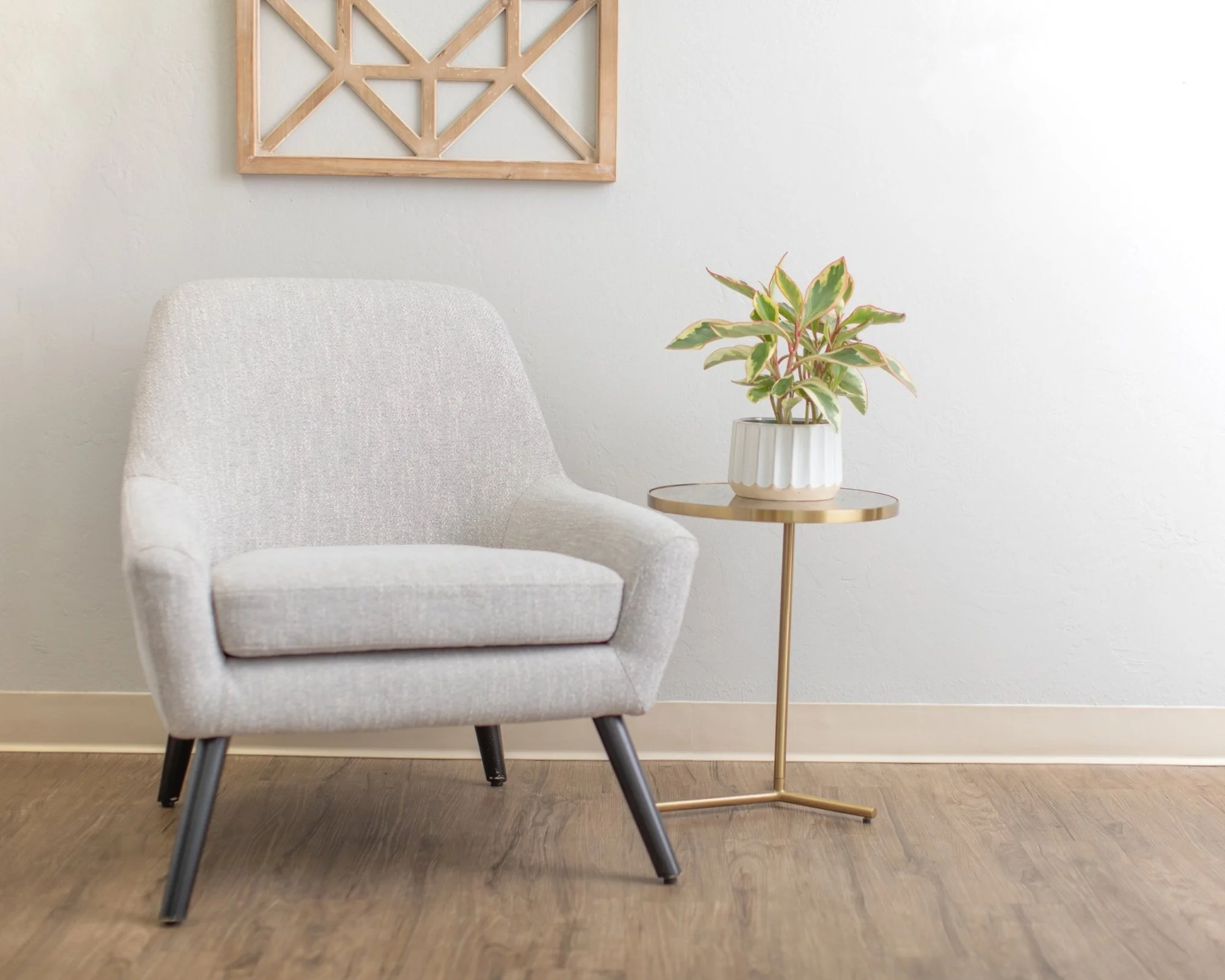 A modern office space corner with a light gray upholstered armchair, a small gold side table with a white potted plant, and a minimalist wooden wall art piece on a white wall. Office waiting space inviting.