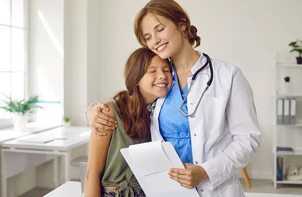 A young girl and a female doctor smiling and hugging in a bright medical office.