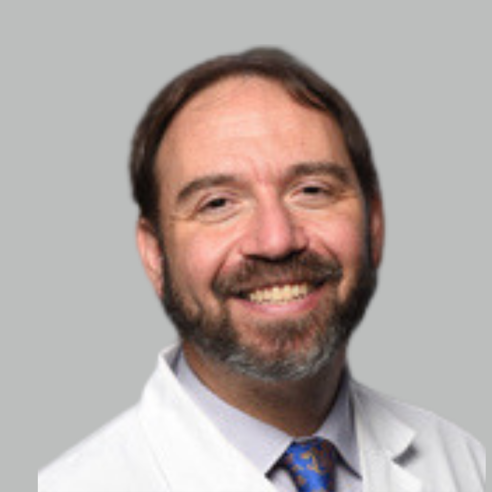 A smiling man with brown hair and facial hair, wearing a white coat, a collared shirt, and a patterned tie, posed against a plain gray background.