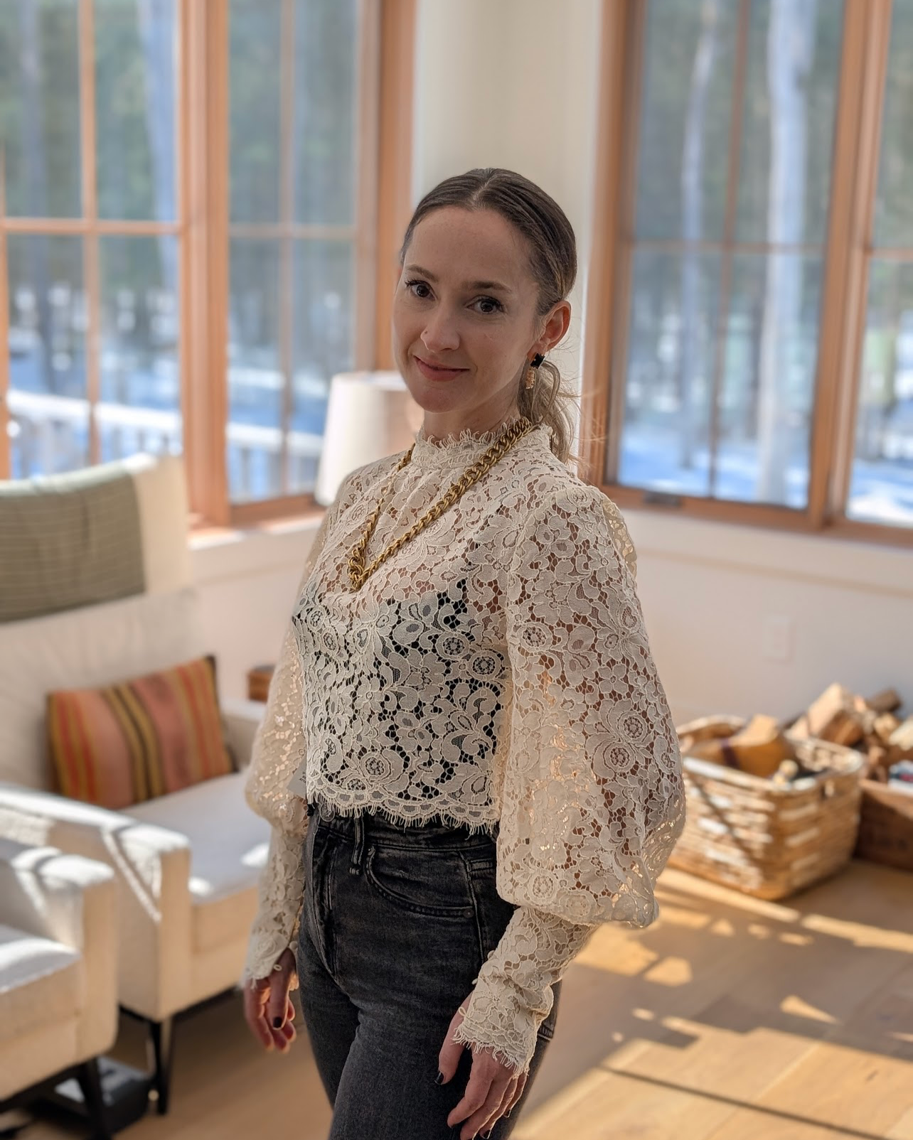 A woman standing indoors near windows with a braided hairstyle, wearing a lace blouse, gold chain necklace, earrings, and dark jeans.