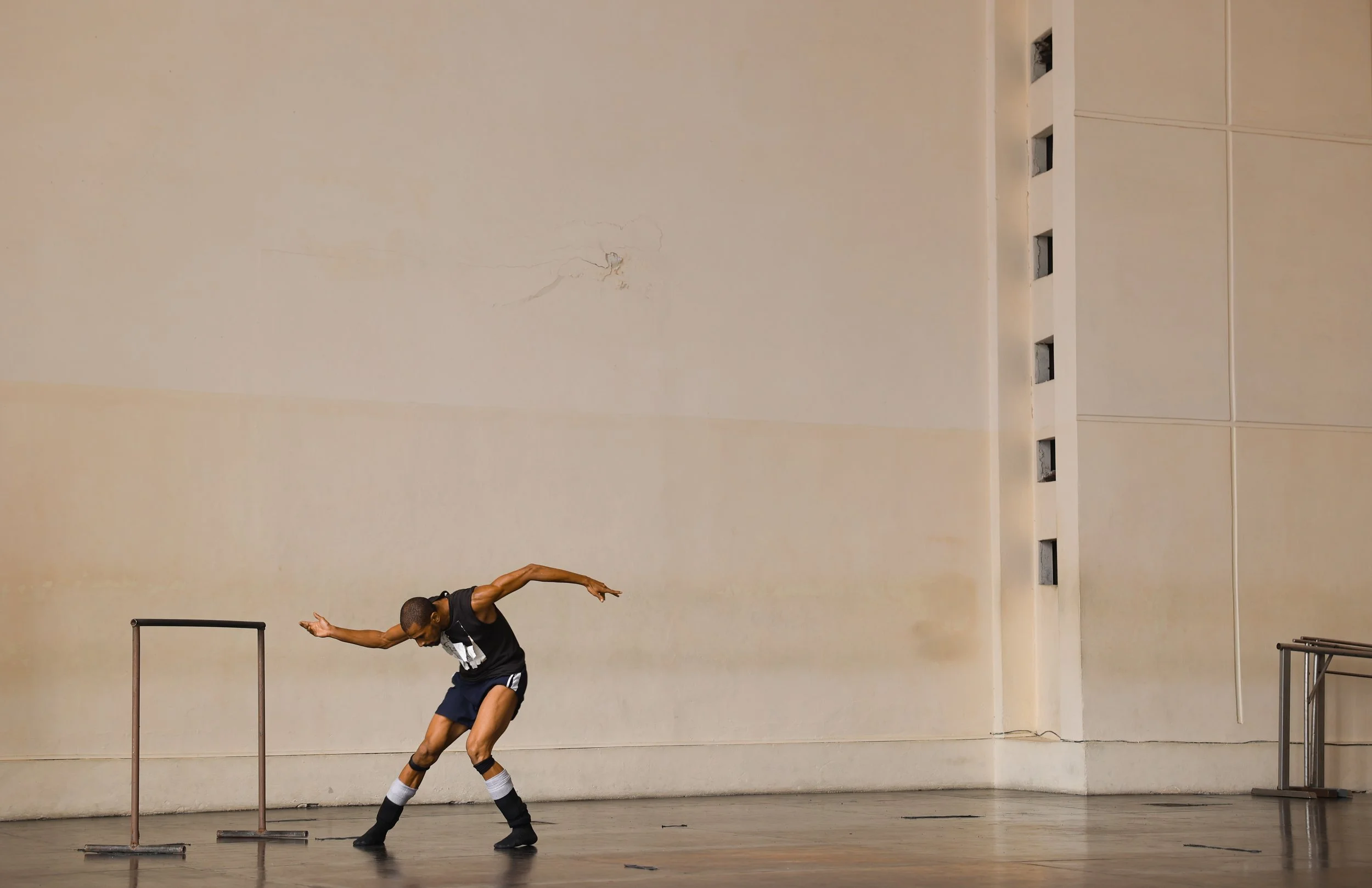 A dancer is performing on stage with a beige wall in the background, some black chairs, and a small set of metal ballet bars. The dancer is spread-eagled, with arms extended to the sides and one leg forward. The dancer wears a black tank top, black shorts, sunglasses, and knee-high black and white socks.