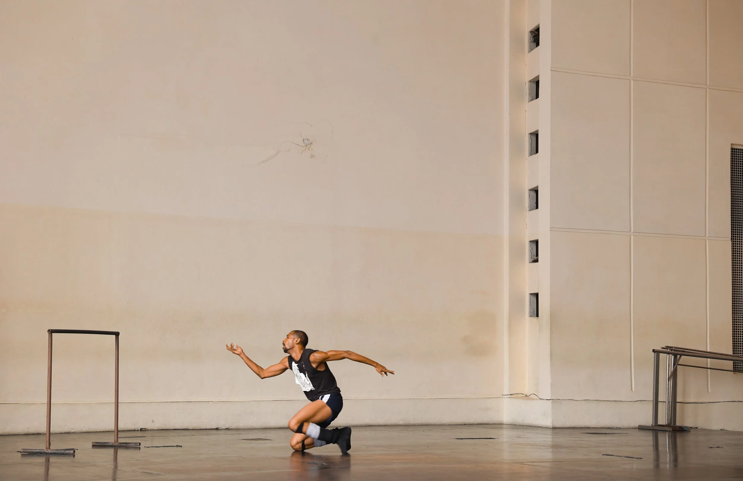 A male dancer is crouching with one knee on the floor and one arm extended, practicing ballet or contemporary dance in a studio with high ceilings, beige walls, and ventilation openings.
