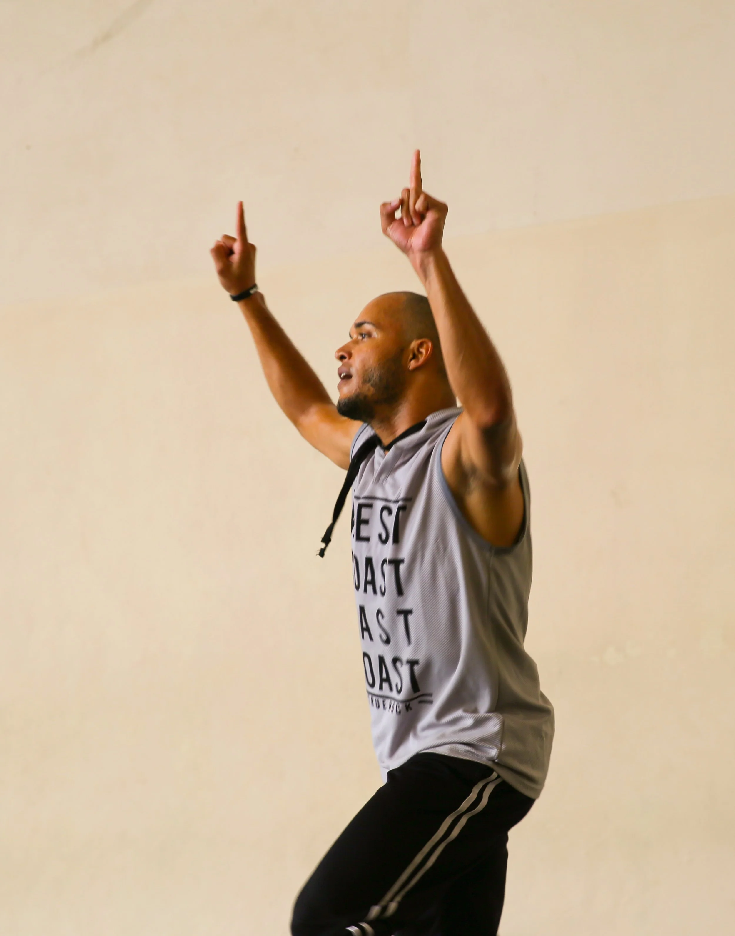 A man with a shaved head and beard, wearing a sleeveless gray shirt with black text, black pants with white stripes, is raising his arms with his index fingers pointing upwards against a plain beige wall.