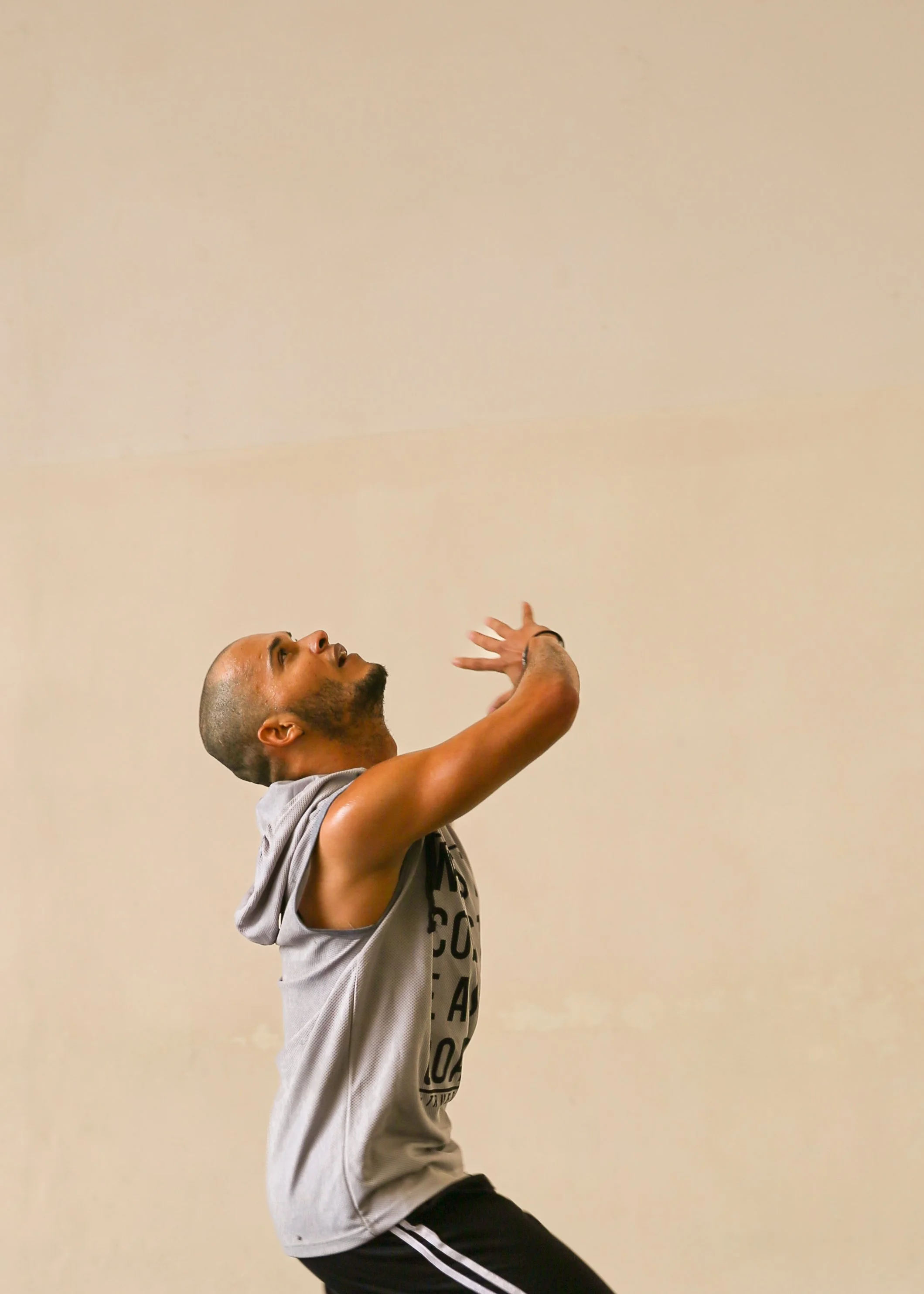 A man in a gray sleeveless hoodie and black and white striped pants performing a dance move against a plain beige wall.