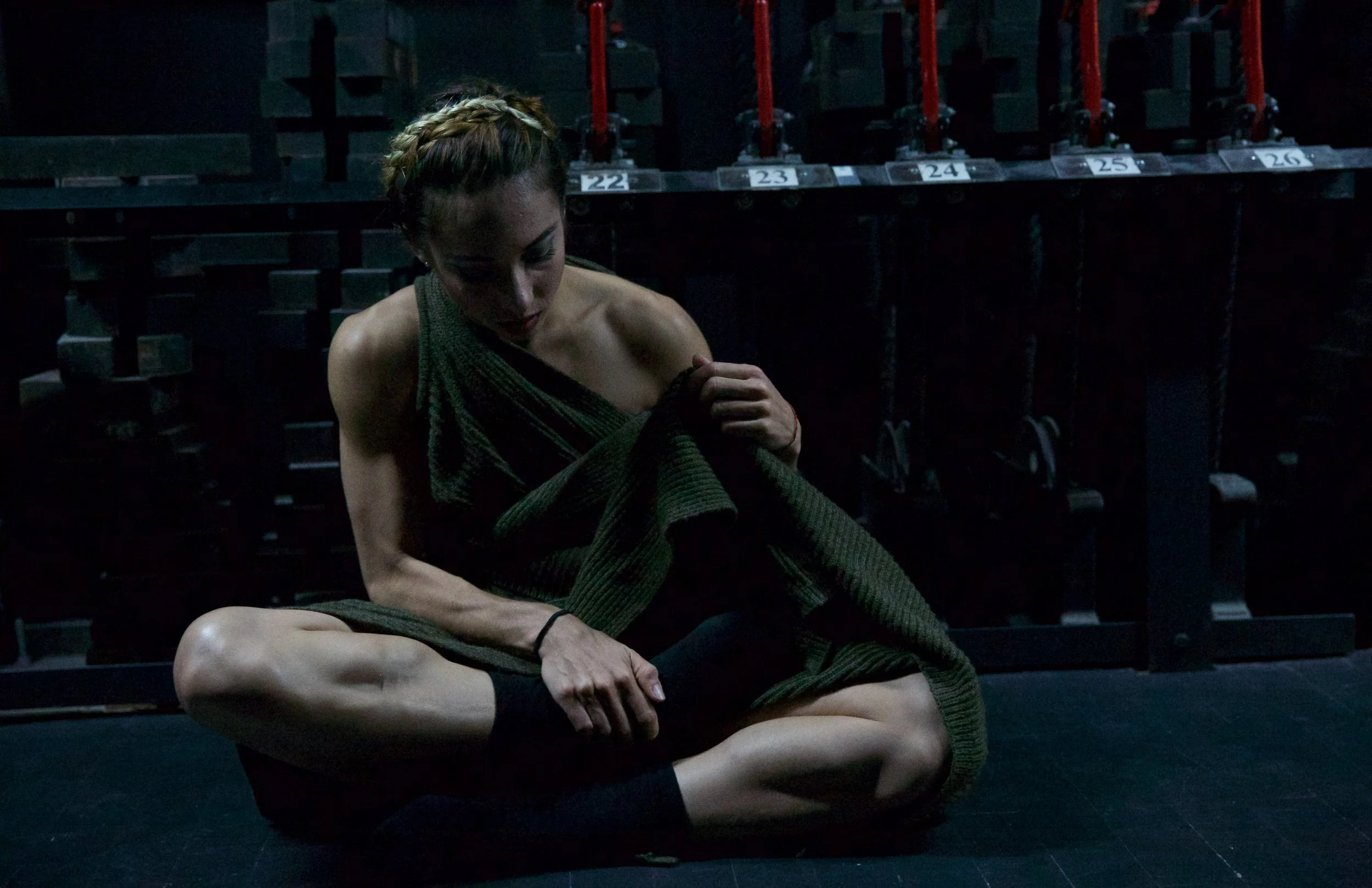 A woman with braided hair sitting cross-legged in a dark room, wrapping herself in a green blanket. The background features black storage shelves with labels numbered 22 to 26 and red pipes.