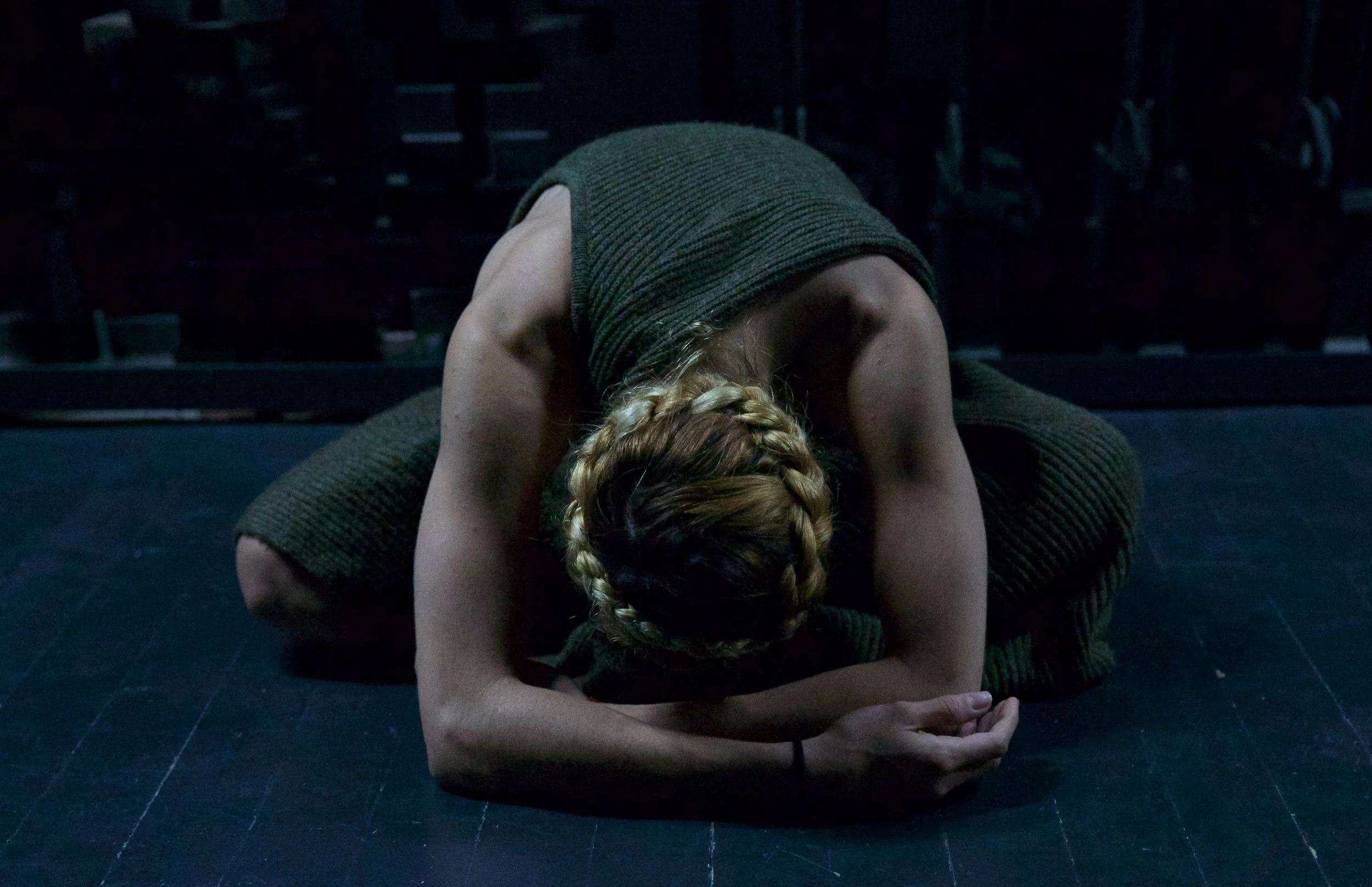 A person with blond hair braided into two pigtails is performing a yoga pose on a dark studio floor, wearing a sleeveless top and pants, with their head and arms resting on the ground.