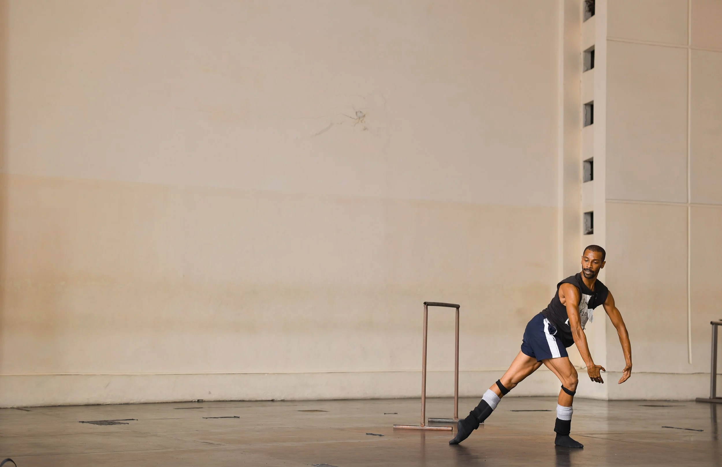 A man in a dance studio performing a stretch or dance move. He is wearing a black tank top, black and white shorts, and black socks and shoes. The studio has a plain beige wall with some damage and a vent on the right side.