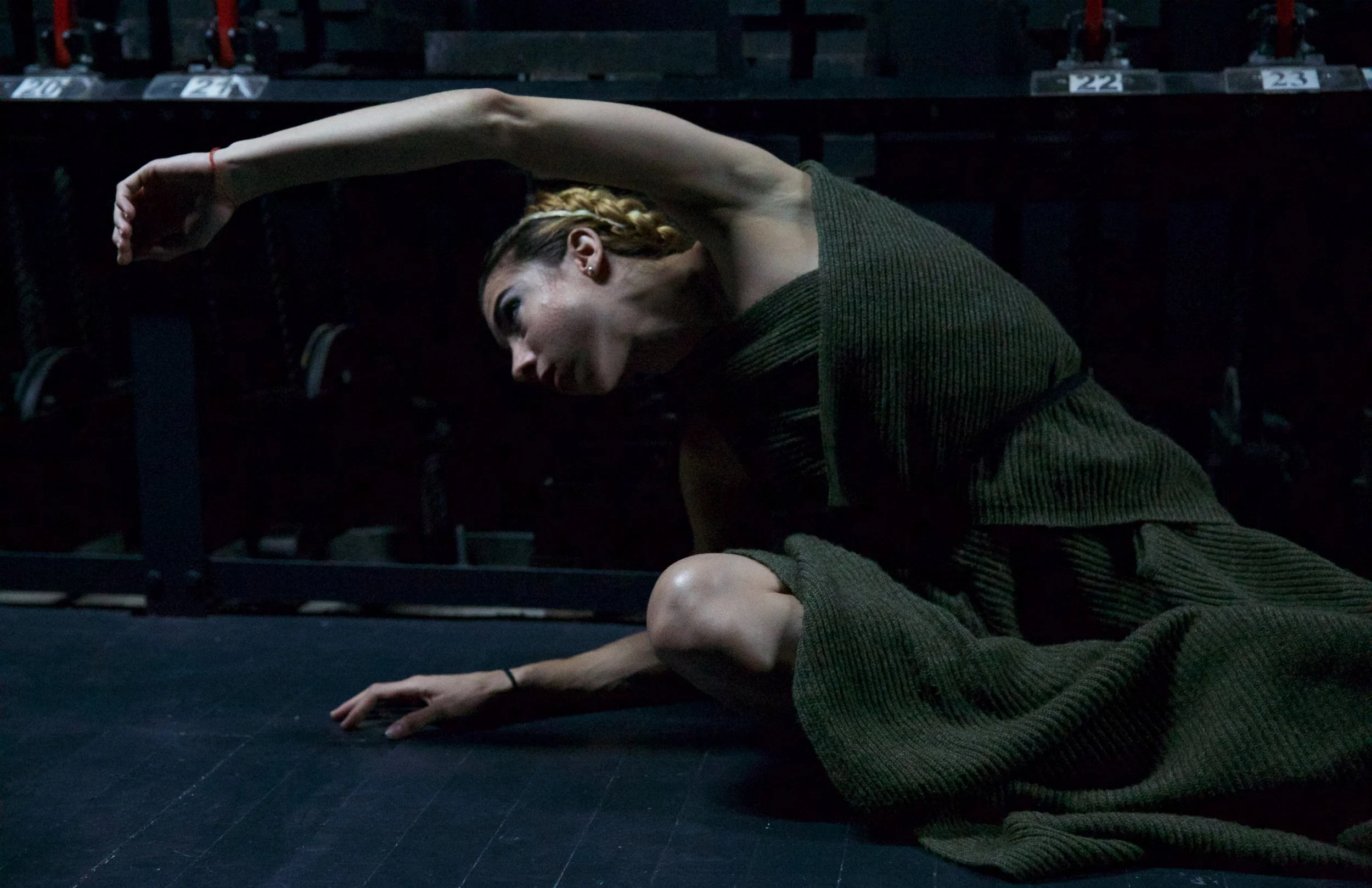A woman with braided hair performs a yoga pose on a dark stage, crouching with one arm extended over her head and the other resting on her knee, with a backdrop of empty chairs.