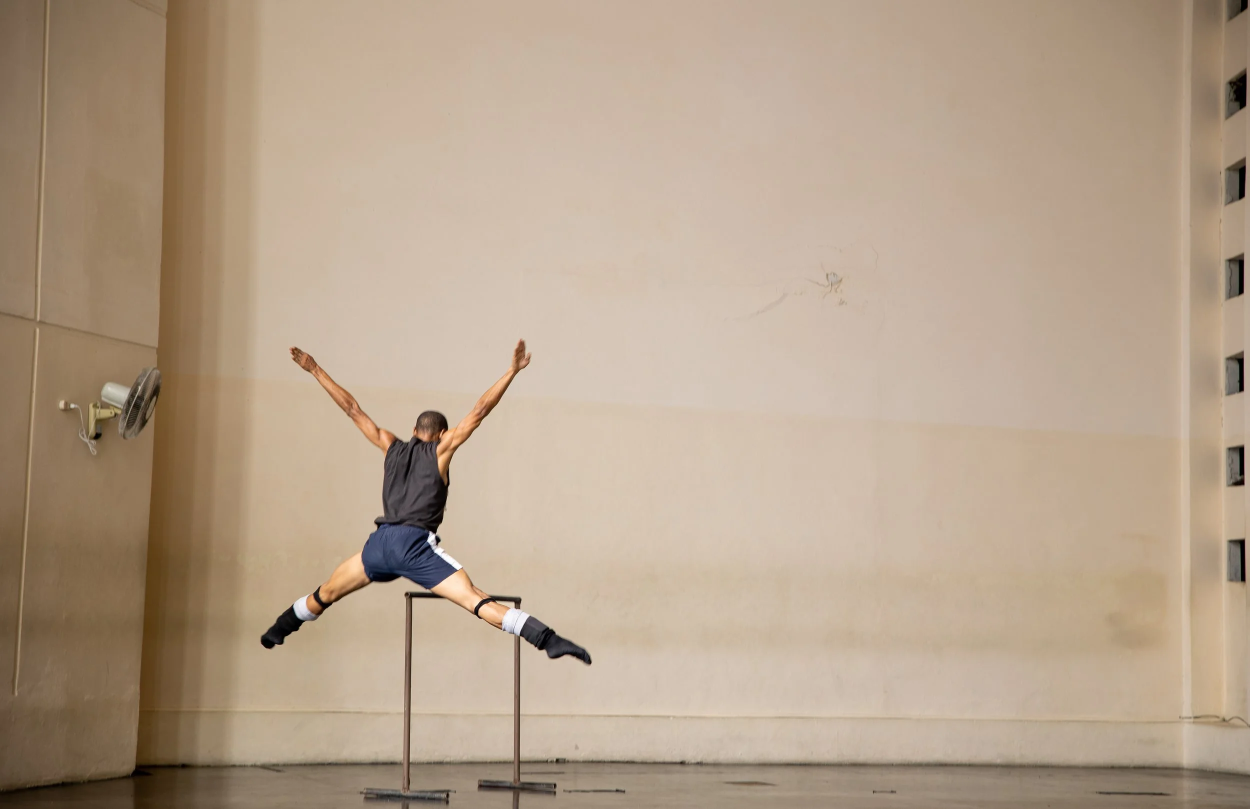 An athlete in a black tank top, shorts, and knee-high socks is jumping over a horizontal pole during an indoor pole vault practice or event. The athlete is mid-air with arms outstretched and legs bent beneath them, against a plain beige wall.