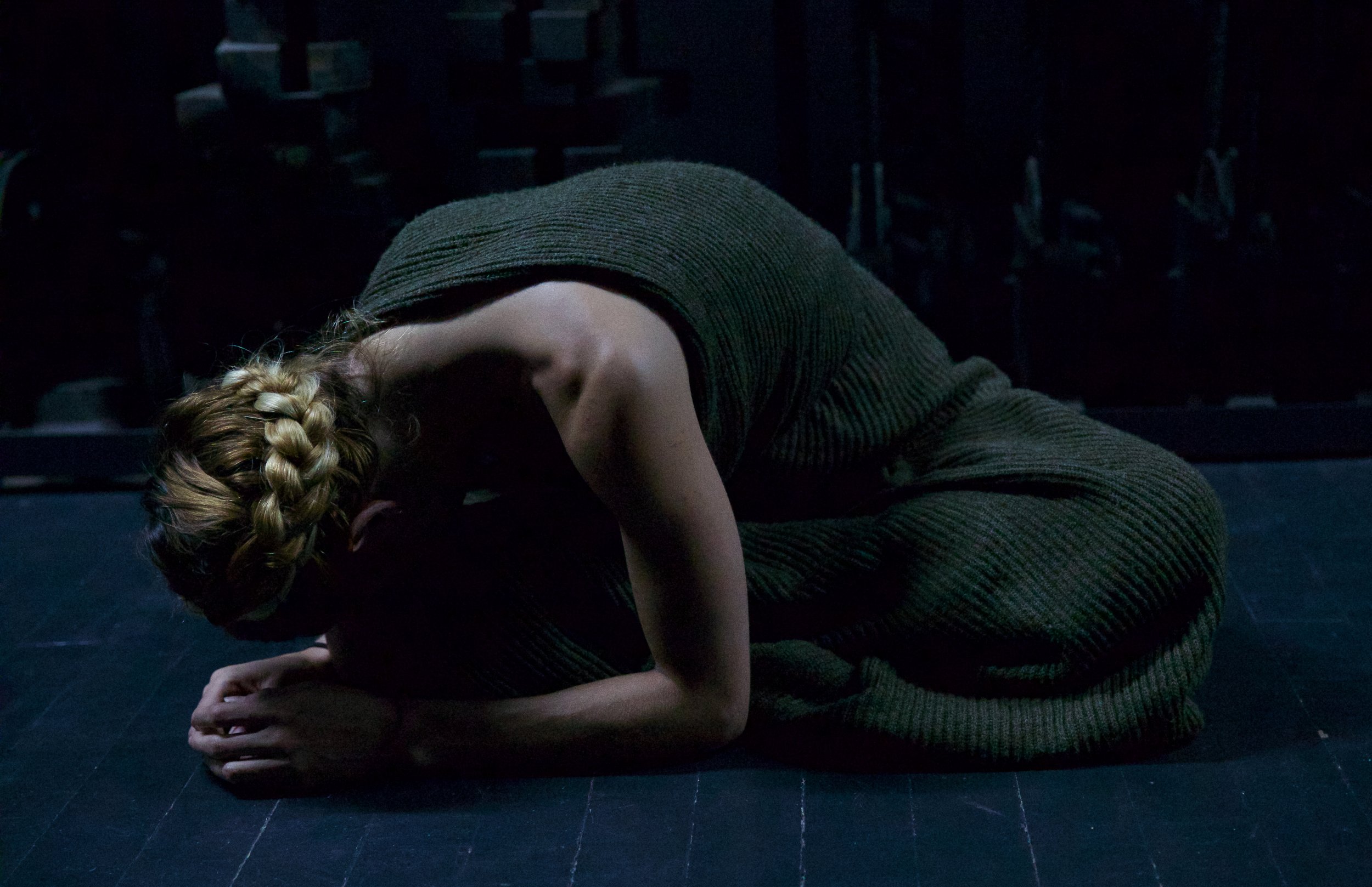 A person with blonde braided hair is practicing yoga on a dark floor, with their upper body bent forward and hands resting on the ground.
