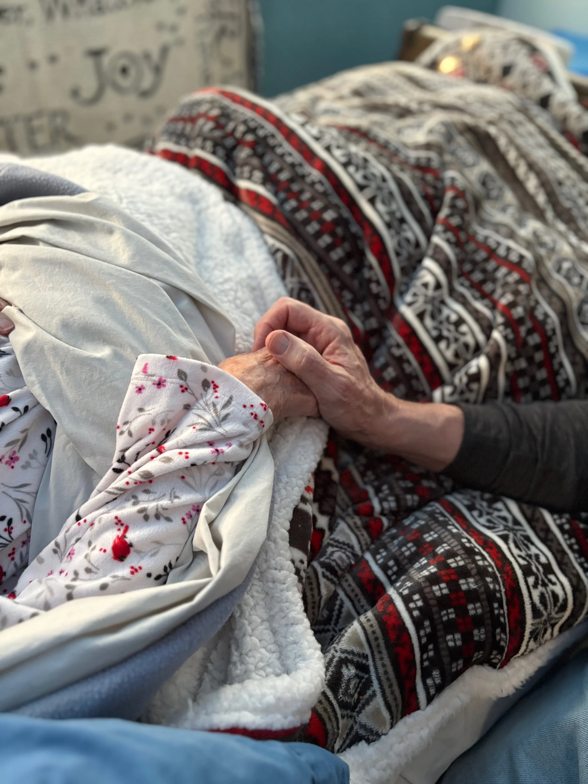 Close-up of hands holding on top of a bedspread. One person is in bed and the other is seated bedside.