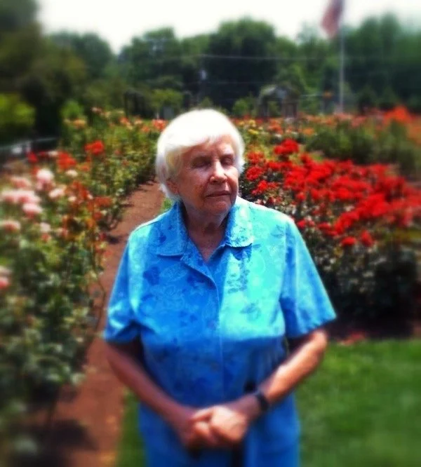 An elderly woman with her eyes closed and hands clasped in front of her, standing in a rose garden on a sunny day.