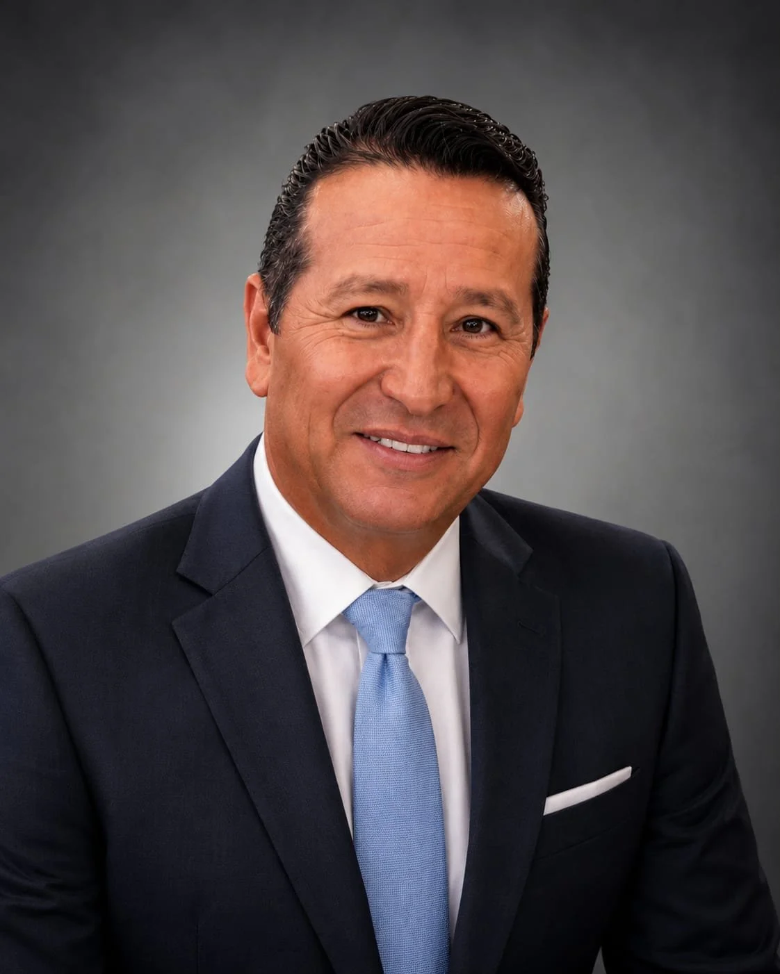 Coach Grimaldo A man in a dark suit, white shirt, and light blue tie smiling at the camera, with a gray background.