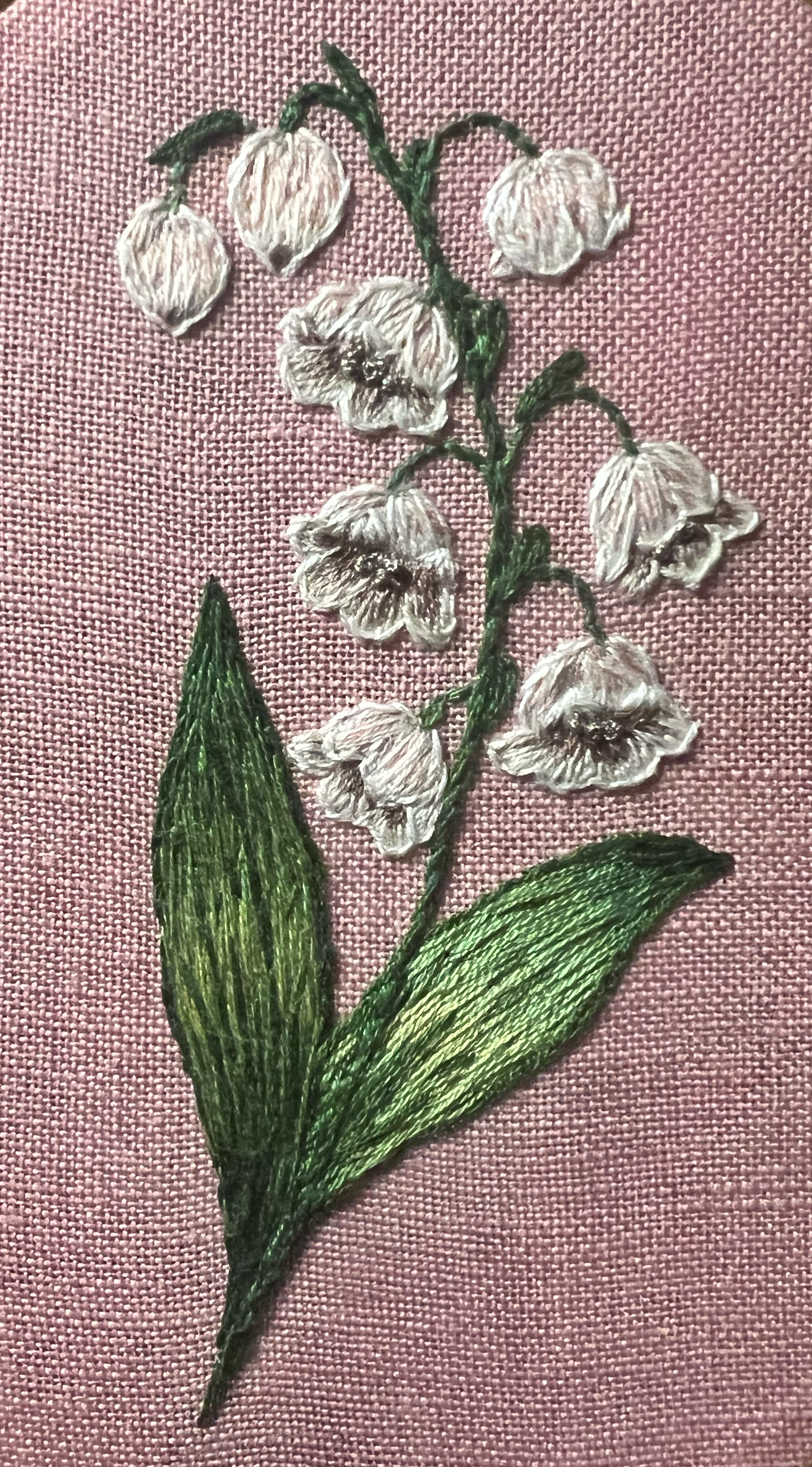 Embroidery of a white flower with green leaves on pink fabric background.