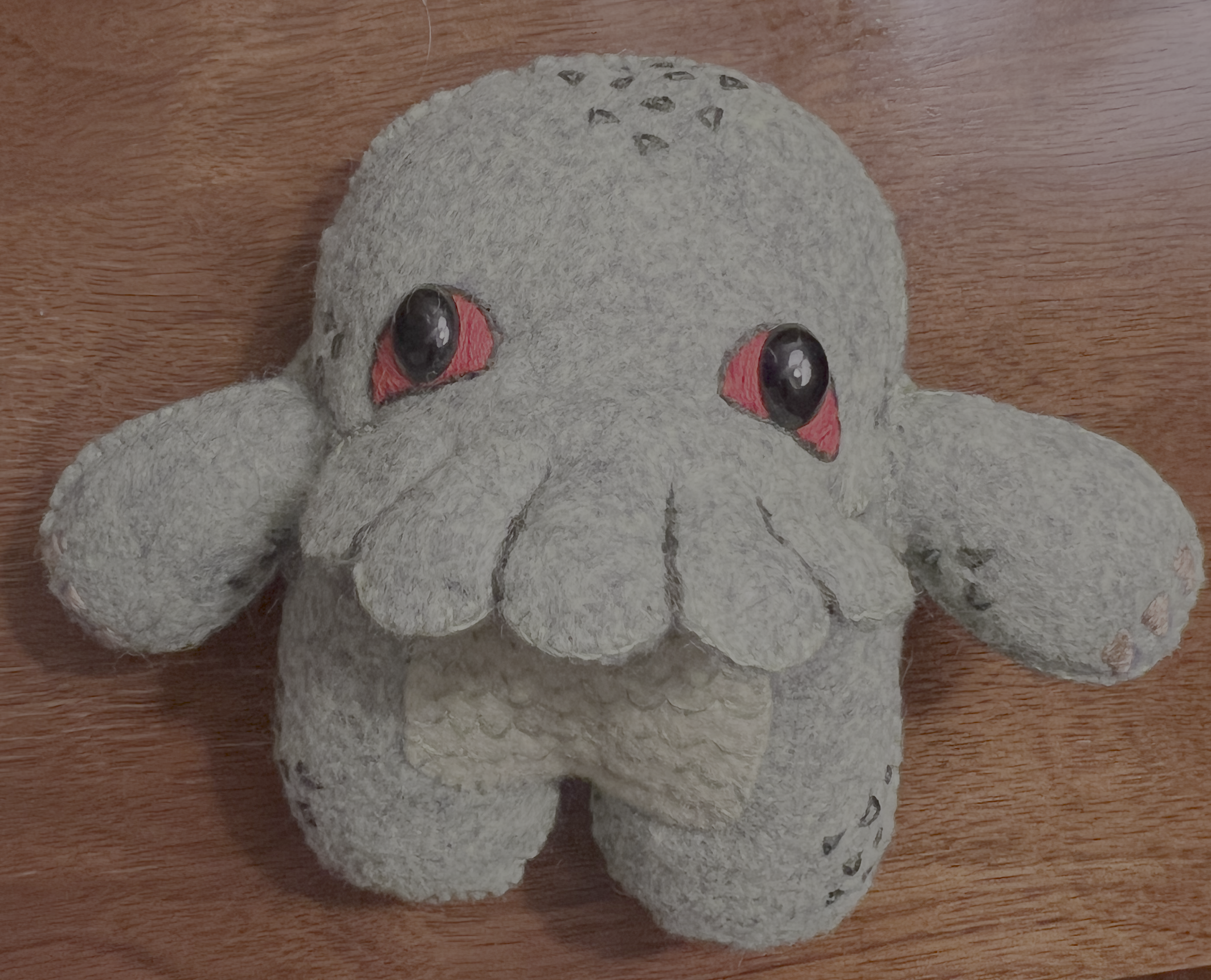 A plush toy of a cute Cthulu with big black eyes and red accents inside the eyes, placed on a wooden surface.