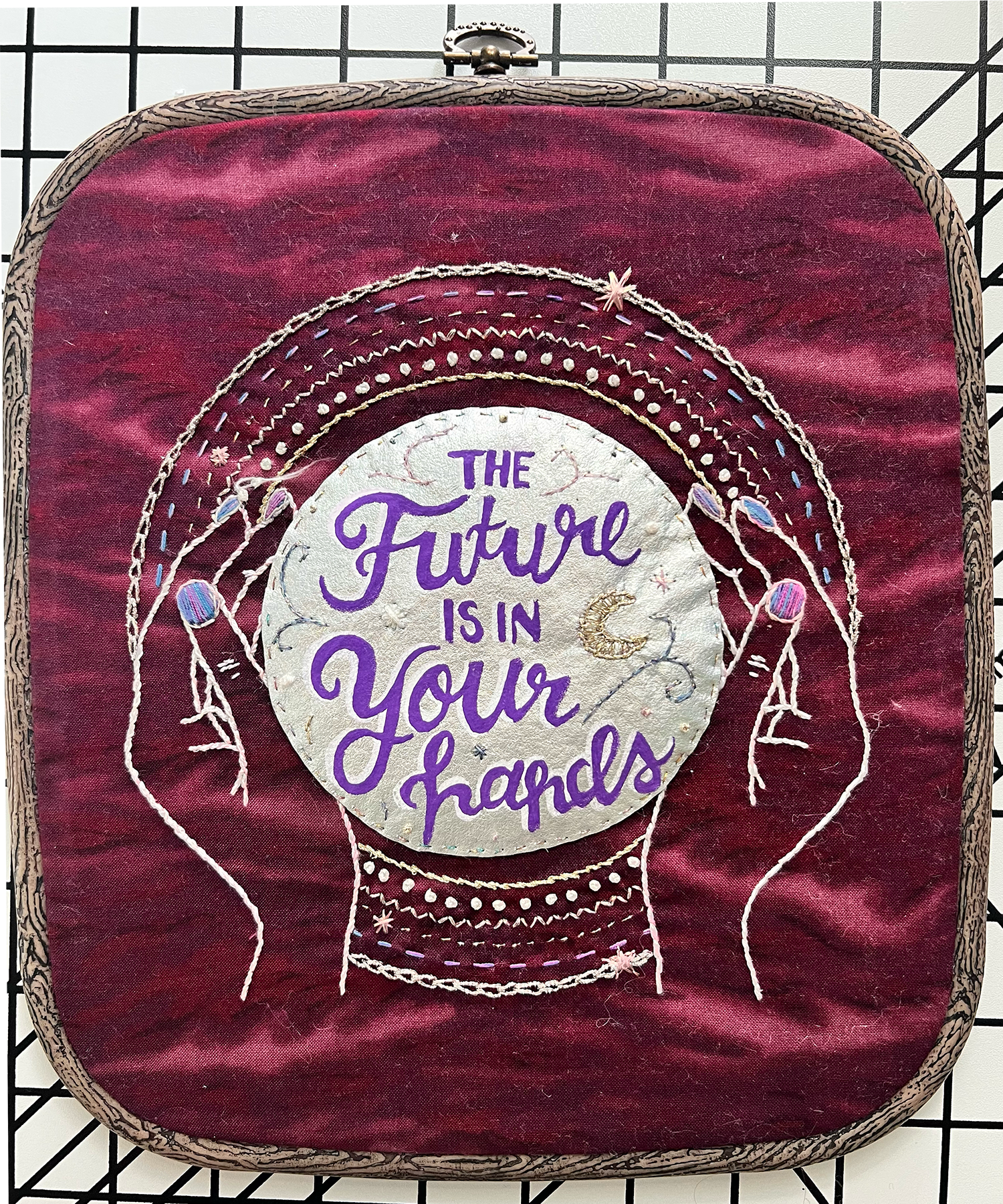 A decorative embroidery on fabric with the quote 'The future is in your hands,' featuring illustrations of hands, stars, and a moon, with colorful thread accents and a velvet background.