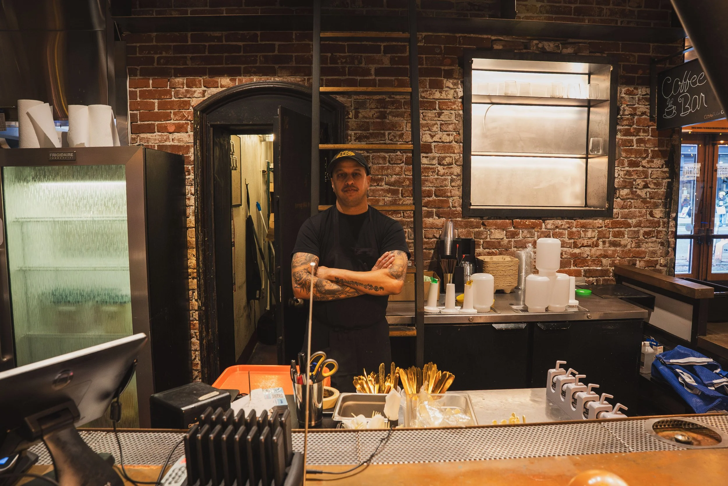 A man wearing a black cap, black shirt, and apron, standing behind a counter in a cafe with exposed brick wall, shelves, and a window. The counter has scoops and containers of utensils.