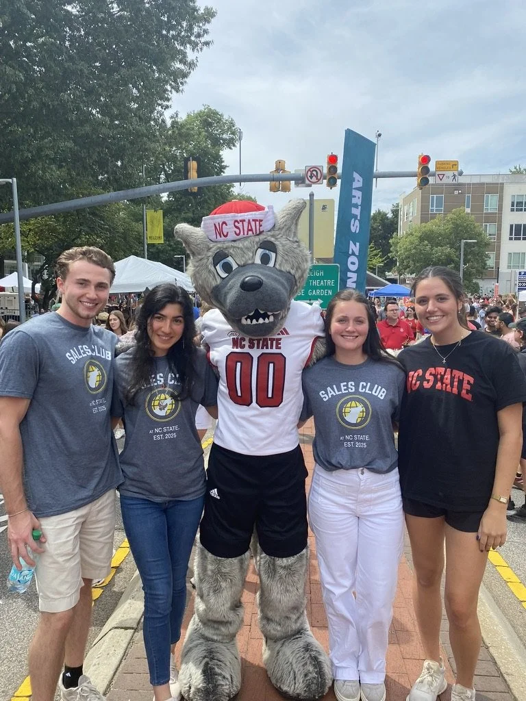 Four young adults standing with a wolf mascot at an outdoor festival. The mascot has gray fur, a black nose, and wears a NC State jersey and hat. The people are wearing NC State themed shirts and are smiling for the photo.