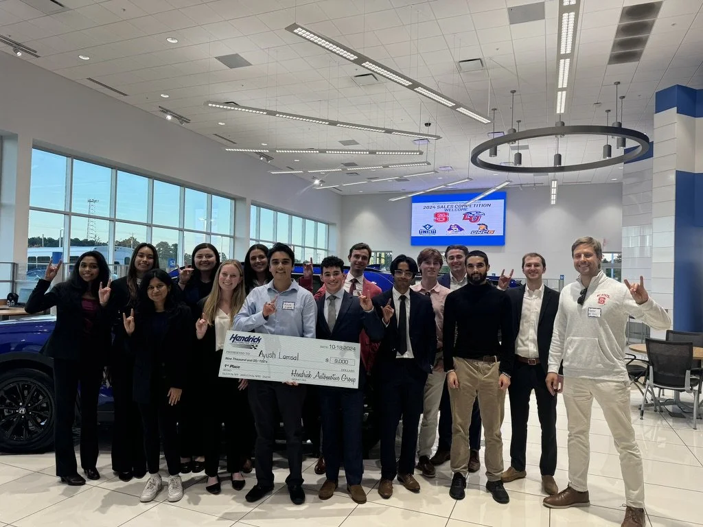 A group of young adults indoors, celebrating a recent achievement, holding a large check, making victory signs, and smiling at the camera.