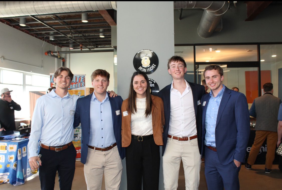 Group of five young adults in business casual attire smiling at a company or career event inside a modern office space.