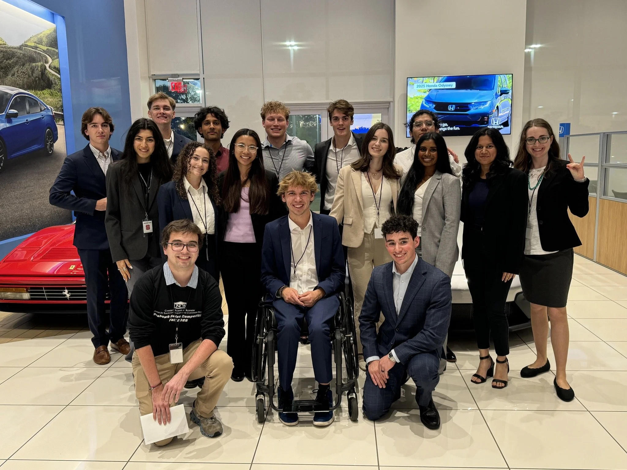 Group of young adults dressed in business attire posing together inside a car dealership showroom with cars and screens in the background.