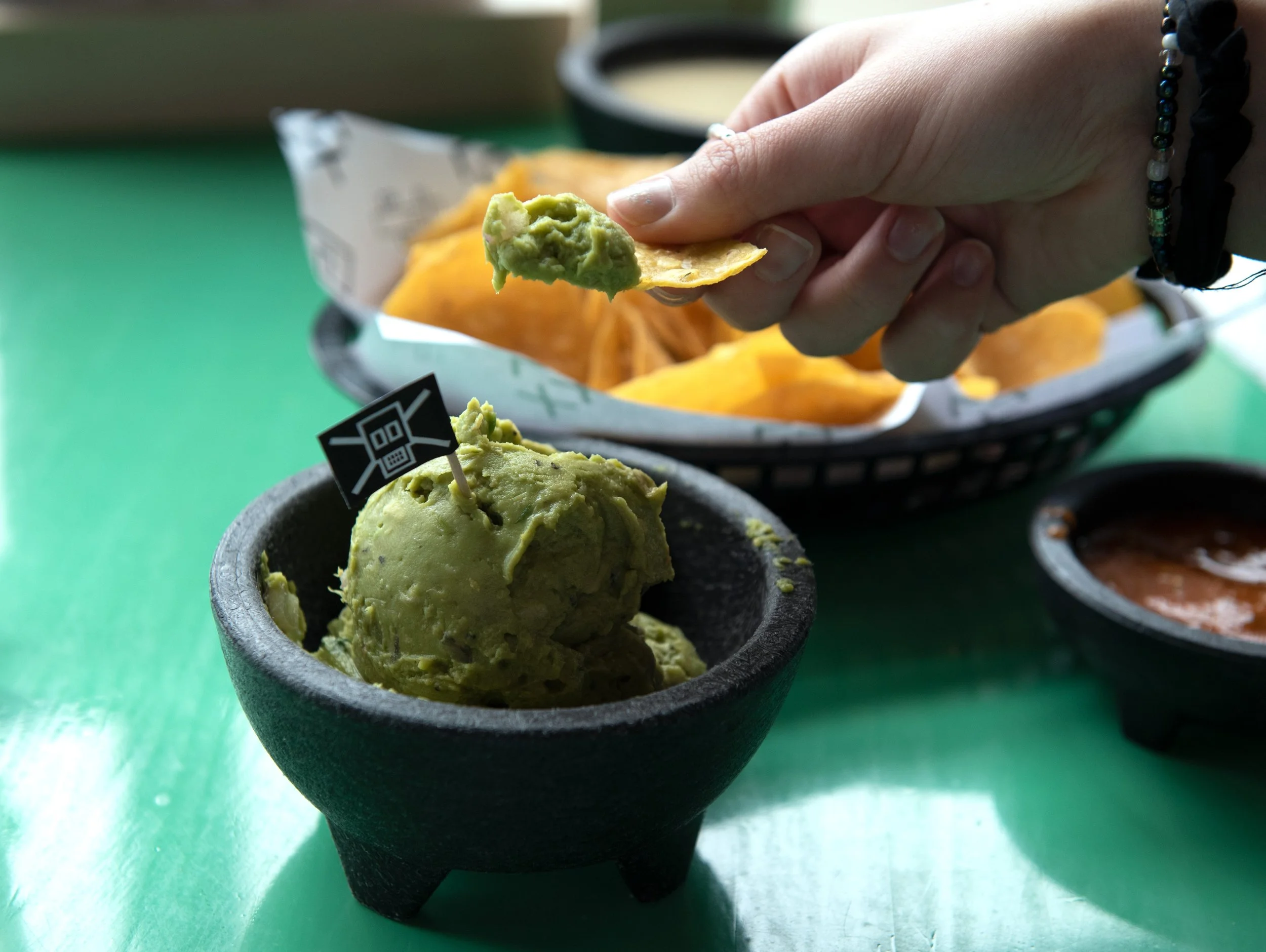 A bowl of guacamole with a small flag on top and a hand dipping a chip into it. Background includes a basket of tortilla chips and a bowl of salsa on a green table.