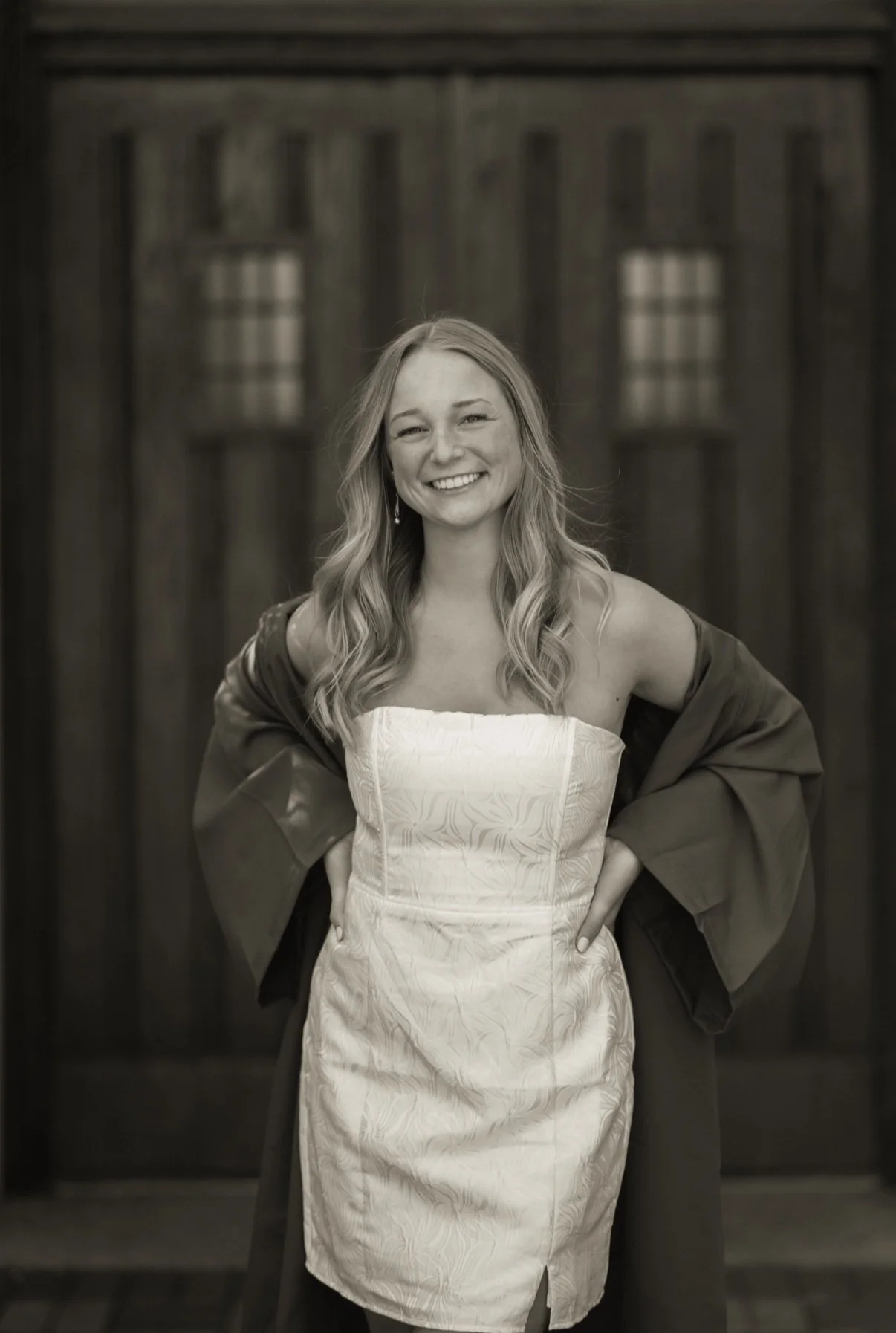 Smiling woman in graduation gown and dress standing in front of wooden door.