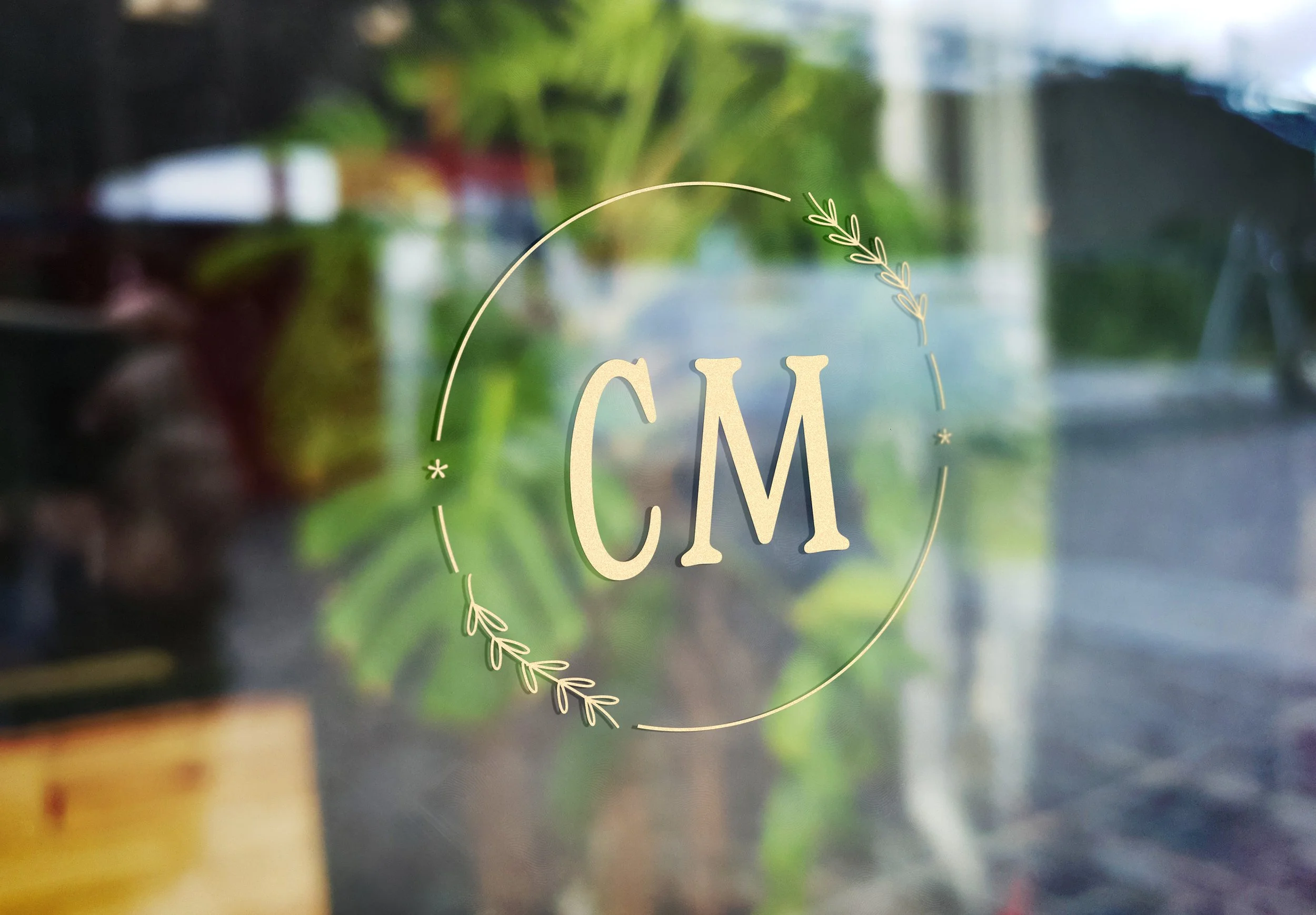 Gold letter logo "CM" inside a circle with decorative leaves on glass surface.