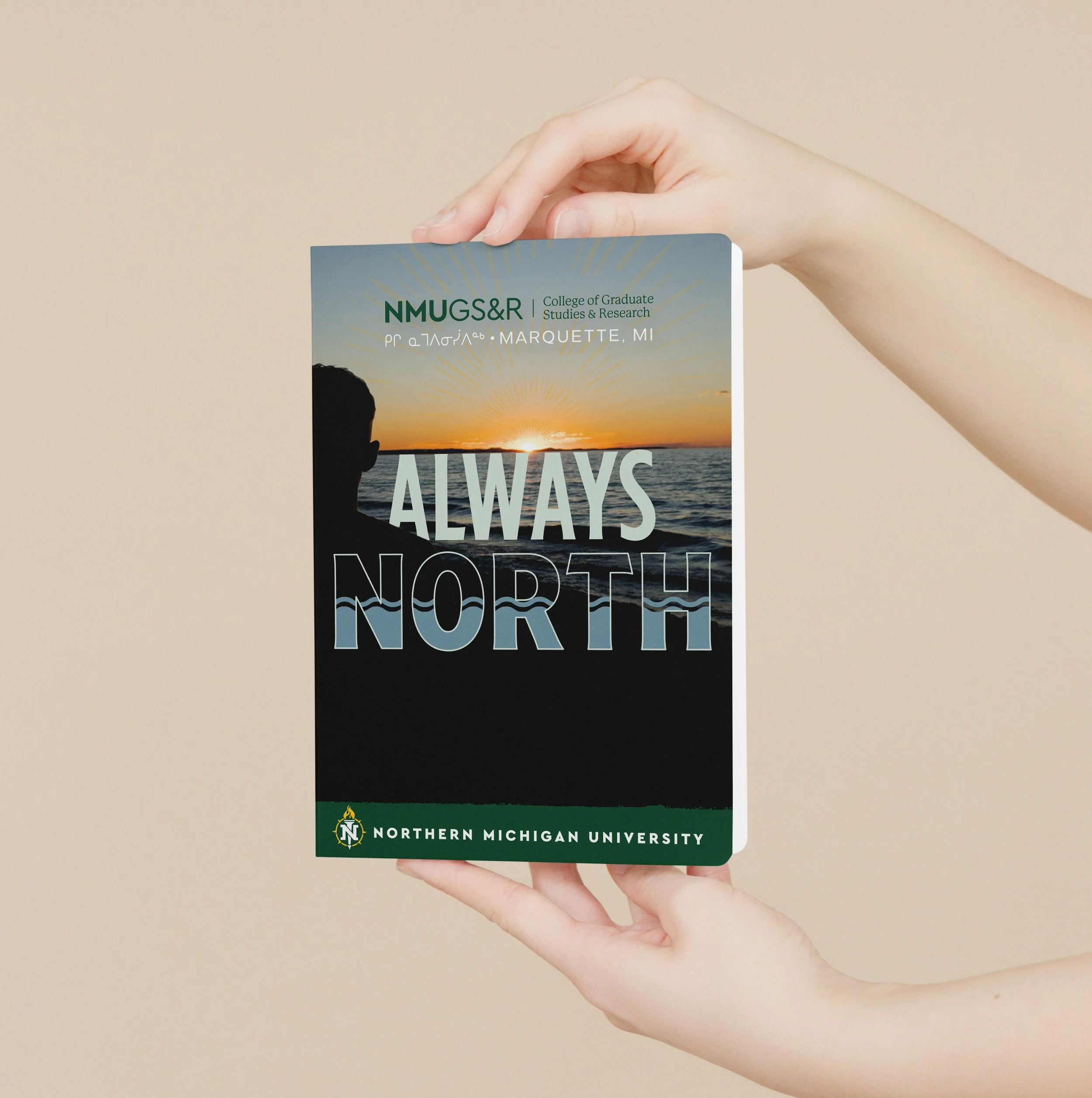 Hands holding a Northern Michigan University poster with "Always North" text, featuring a silhouette against a sunset over water, promoting the College of Graduate Studies and Research in Marquette, MI.