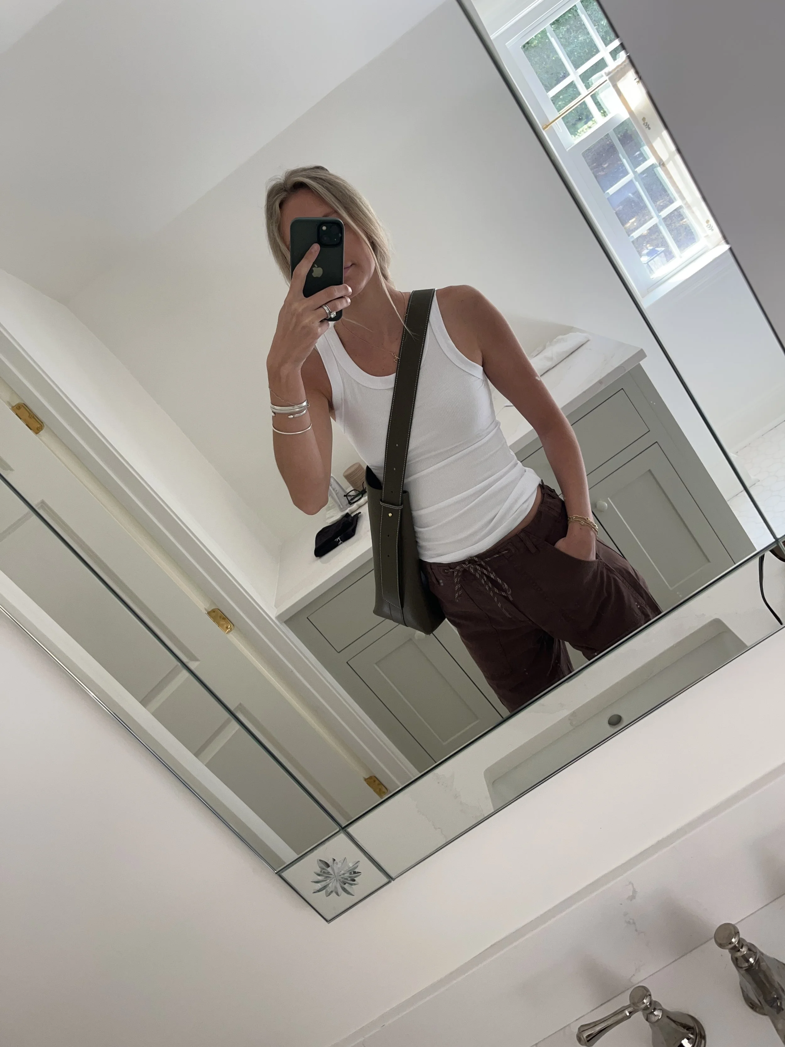 A woman taking a mirror selfie in a spacious living room with wooden floors, large windows, and minimal furniture, wearing a beige jacket, black top, blue jeans, and tan shoes.