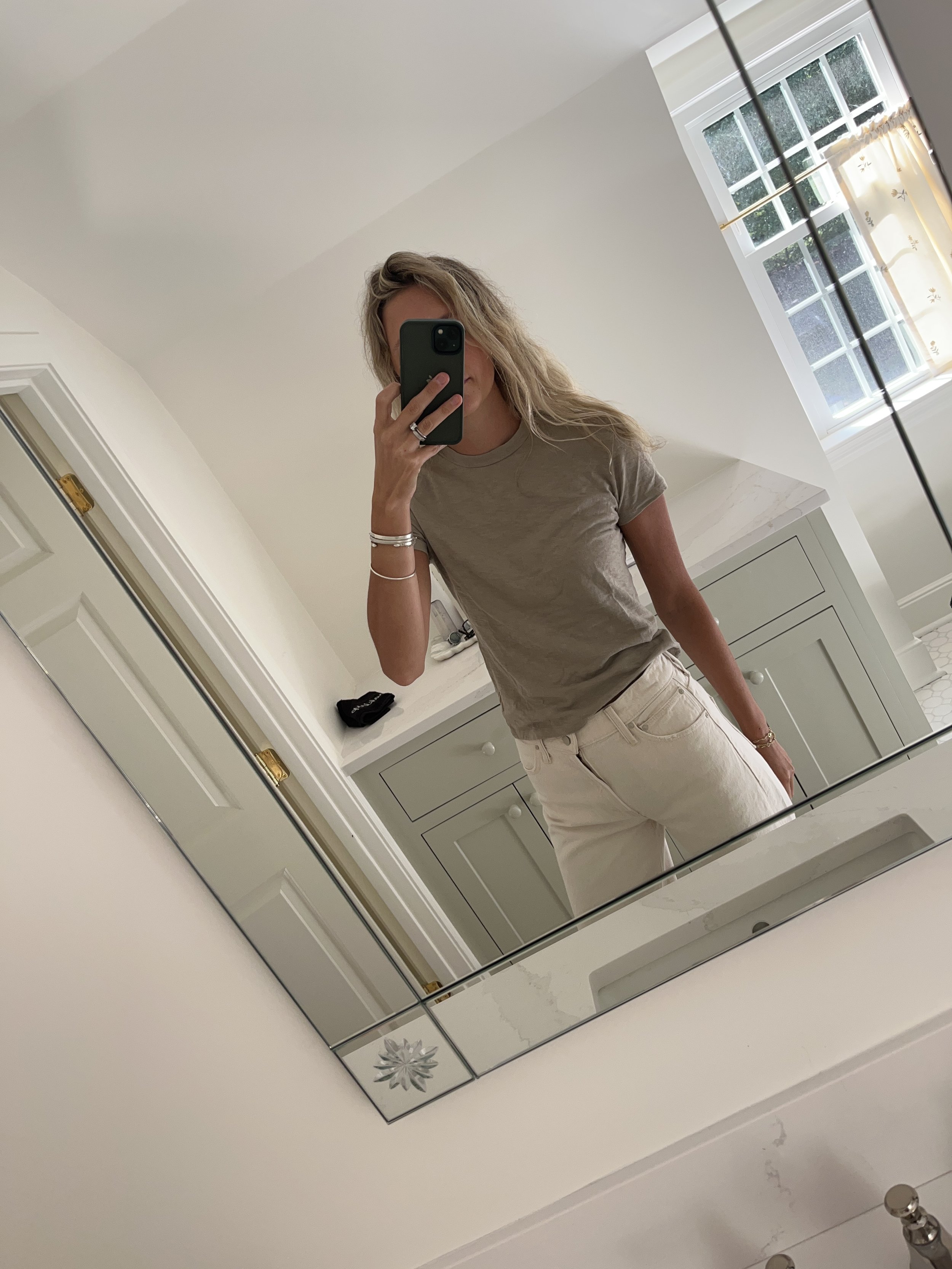 A woman taking a mirror selfie in a spacious living room with wooden floors, large windows, and minimal furniture, wearing a beige jacket, black top, blue jeans, and tan shoes.