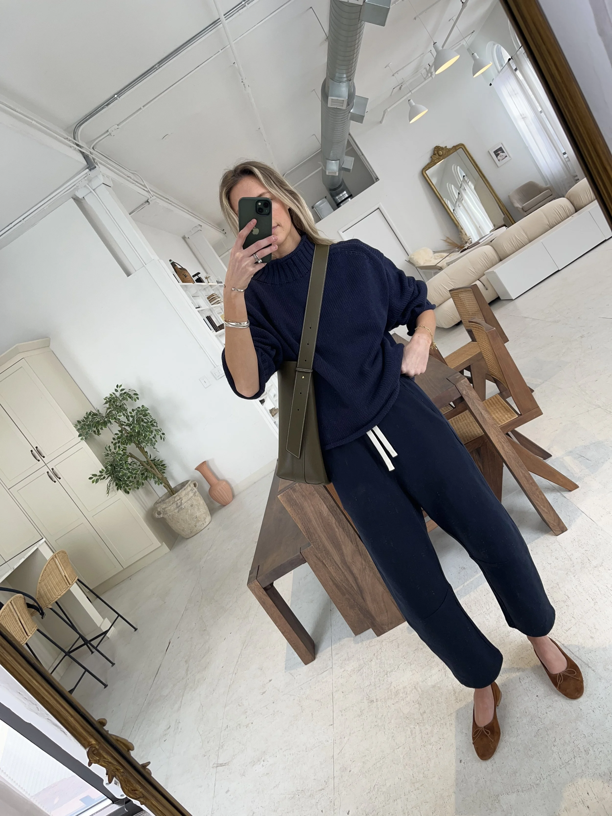 A woman taking a mirror selfie in a spacious living room with wooden floors, large windows, and minimal furniture, wearing a beige jacket, black top, blue jeans, and tan shoes.