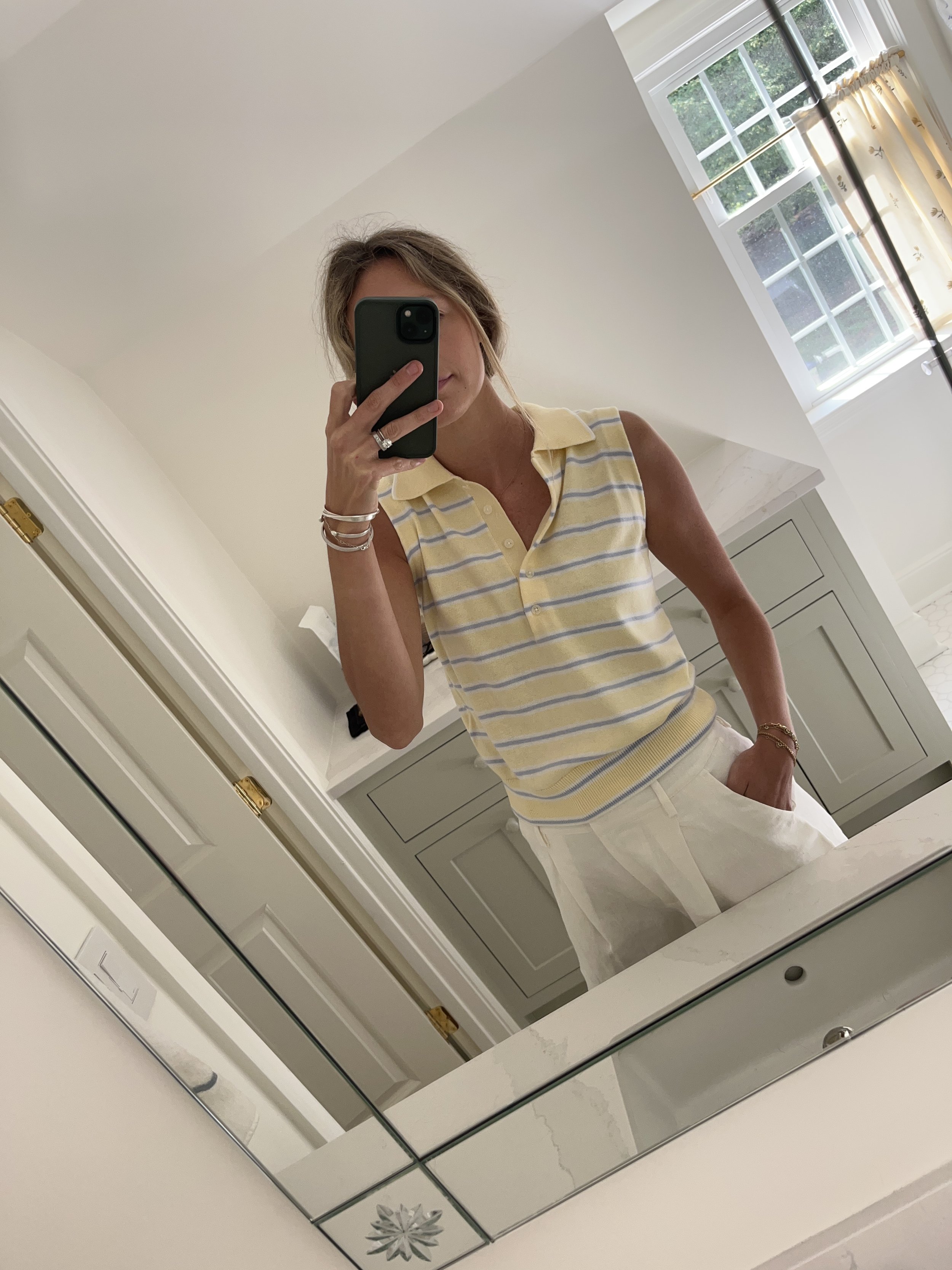 A woman taking a mirror selfie in a spacious, well-lit living room with hardwood floors, large windows, a white sofa, and a side table with a plant.