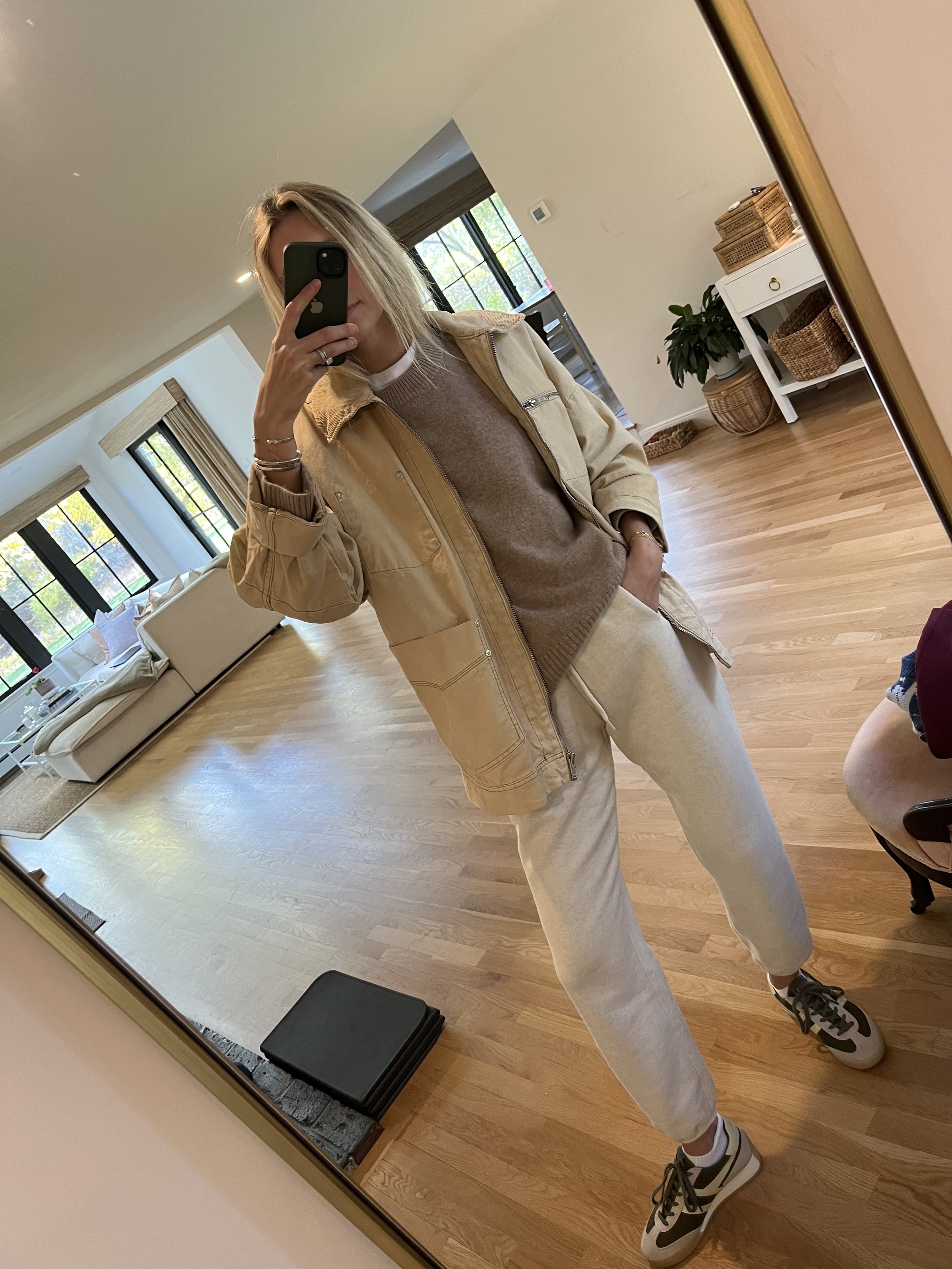 Woman taking a mirror selfie in a spacious living room with hardwood floors, large windows, a white sofa, and a white console table with baskets and a plant.