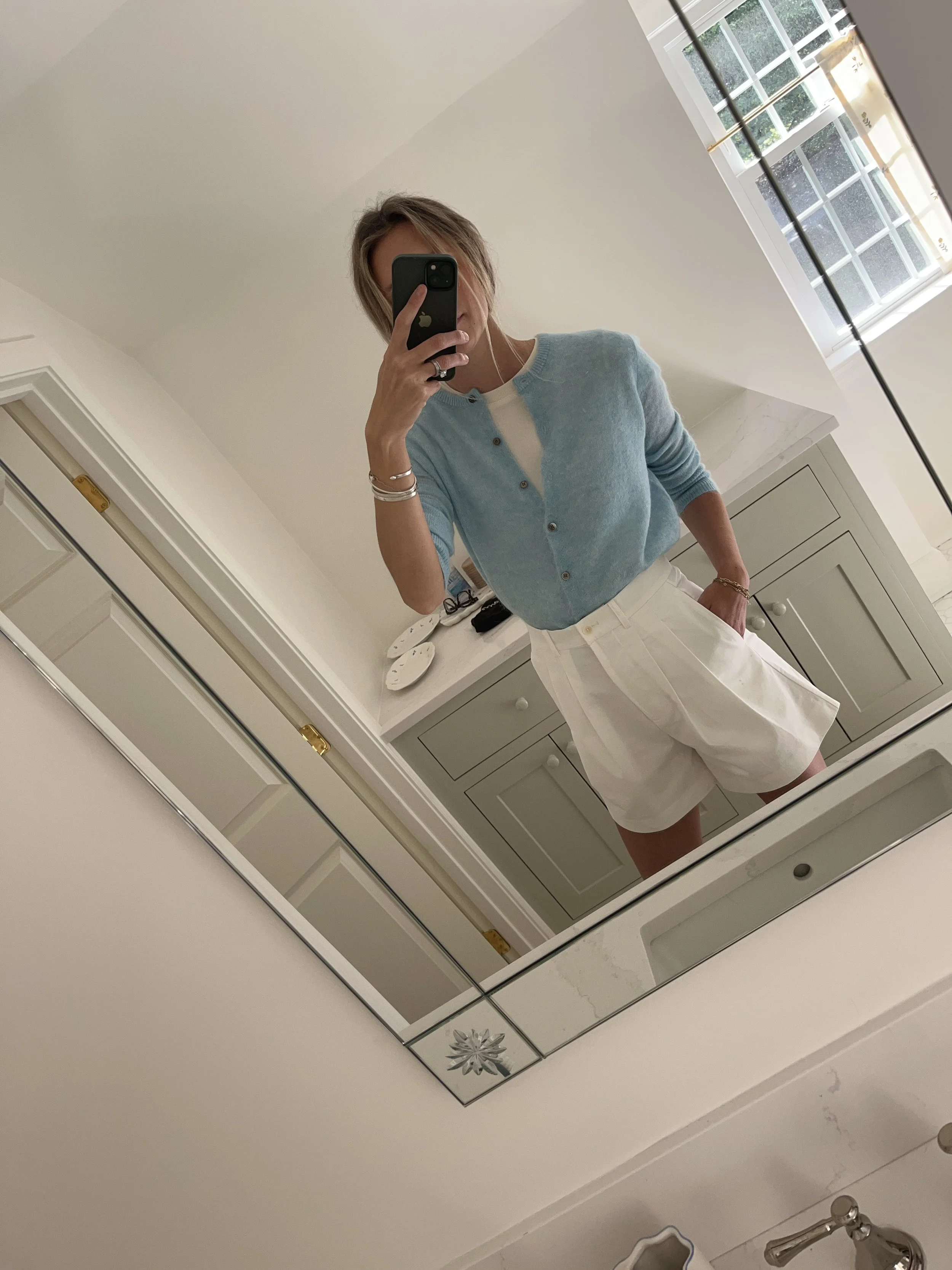 Person taking a mirror selfie in a living room with hardwood floors, wearing a black fleece jacket, gray shirt, black leggings, gray sneakers, and a white cap with a logo.