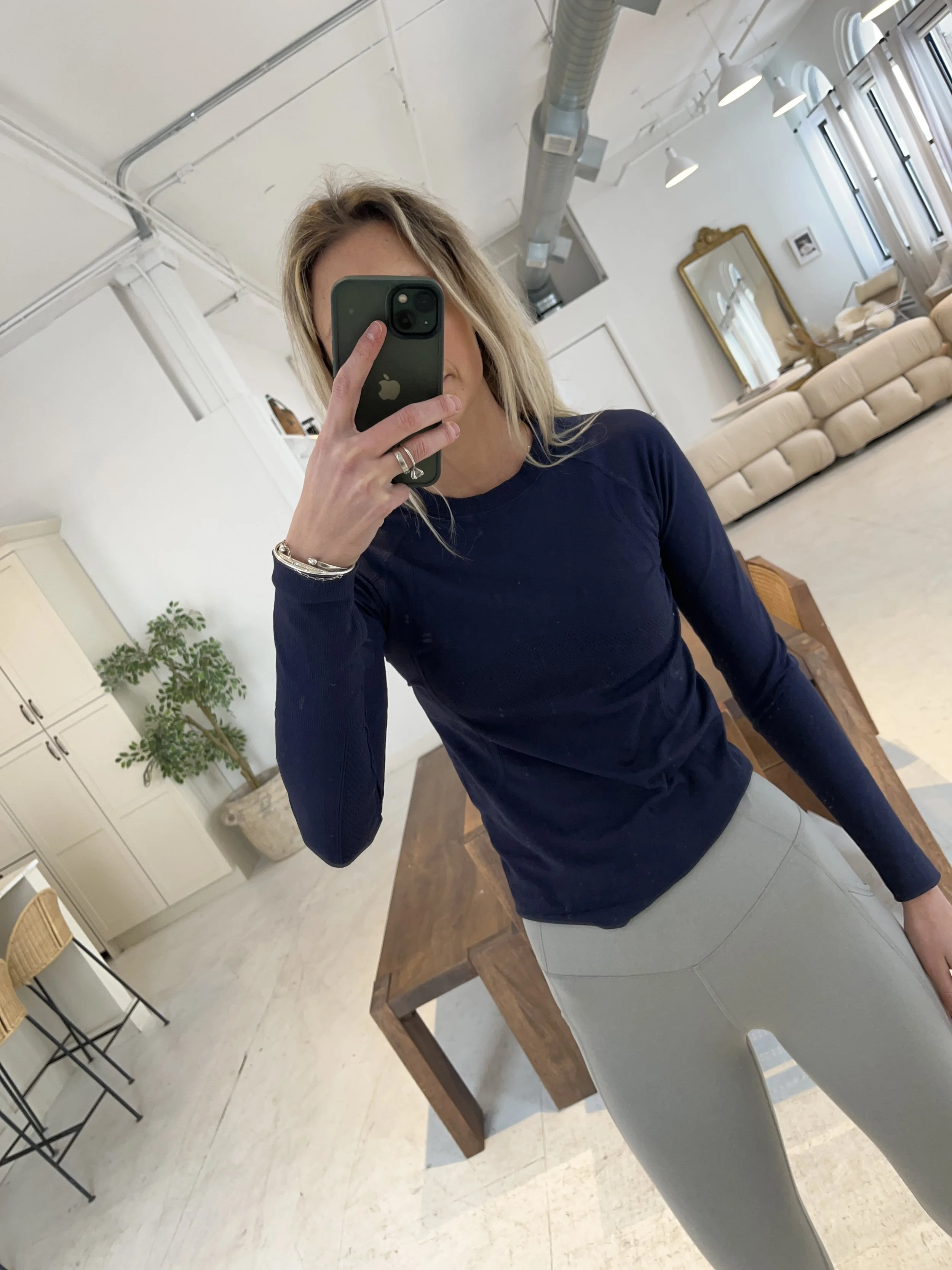 Person taking a mirror selfie in a room with wooden floors, wearing a striped brown and white long-sleeve shirt, light blue jeans, and black and white sneakers.