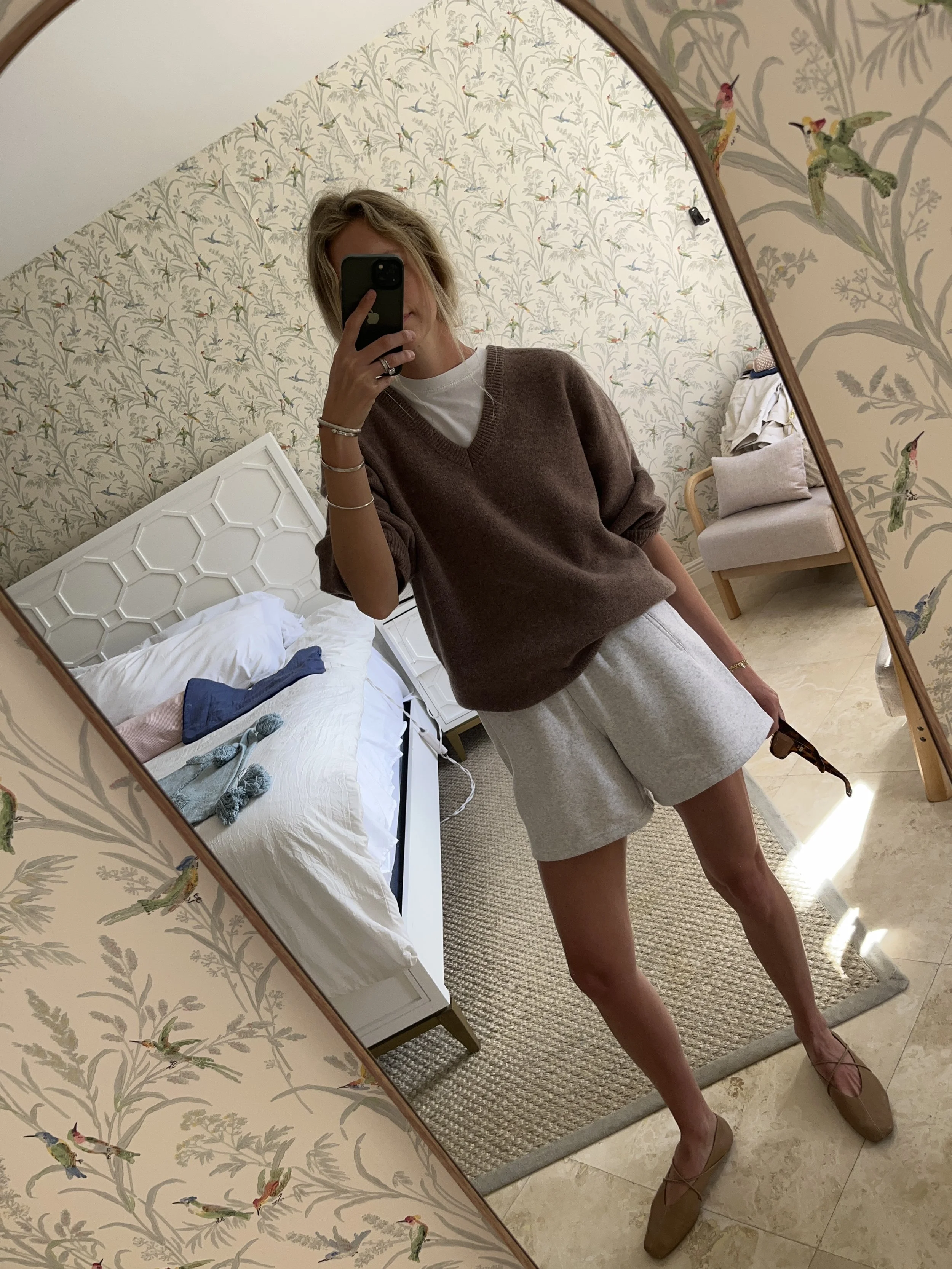 A woman taking a mirror selfie in a spacious living room with wooden floors, large windows, and minimal furniture, wearing a beige jacket, black top, blue jeans, and tan shoes.