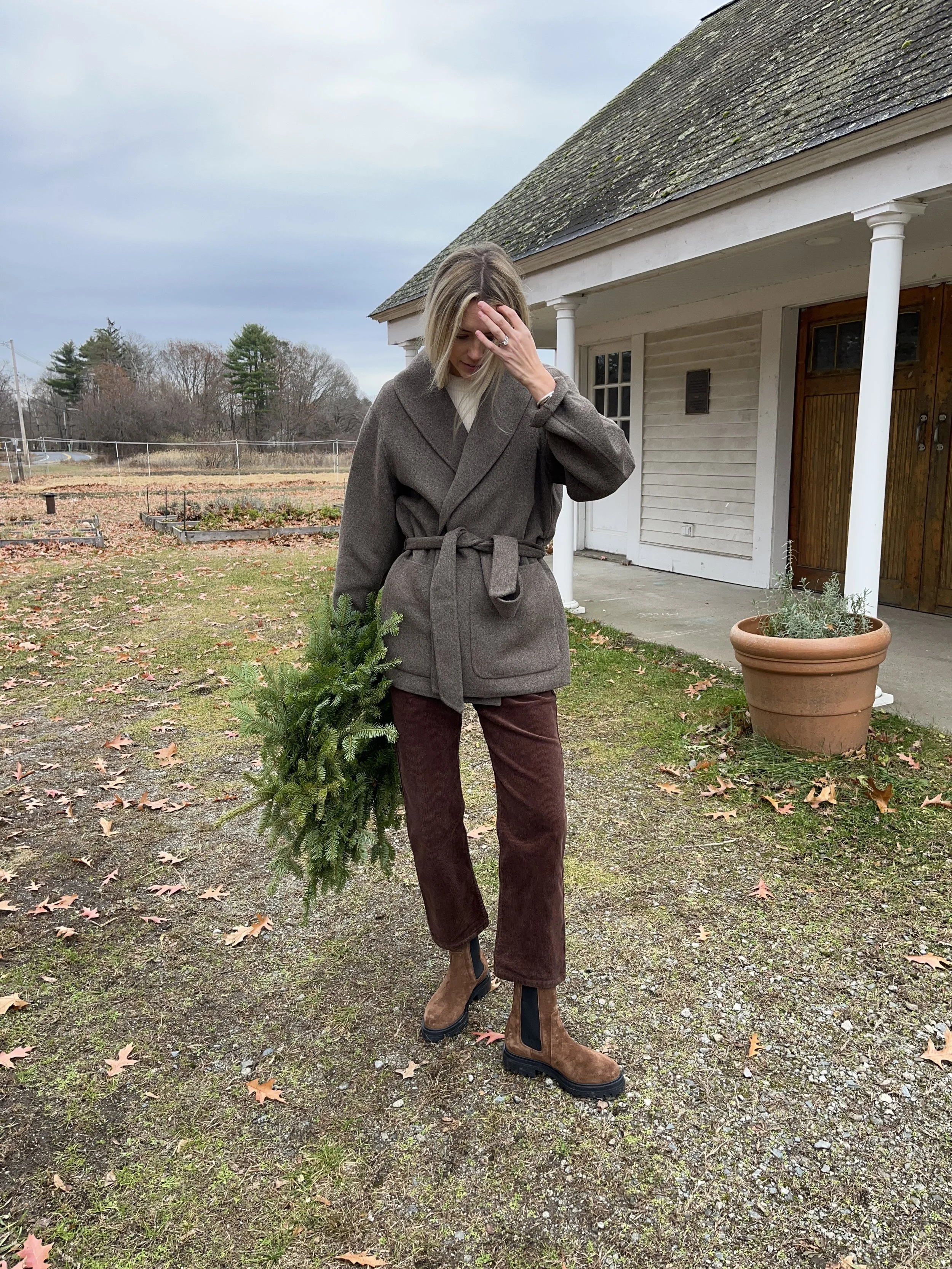 Person taking a mirror selfie in a living room with hardwood floors, wearing a black fleece jacket, gray shirt, black leggings, gray sneakers, and a white cap with a logo.