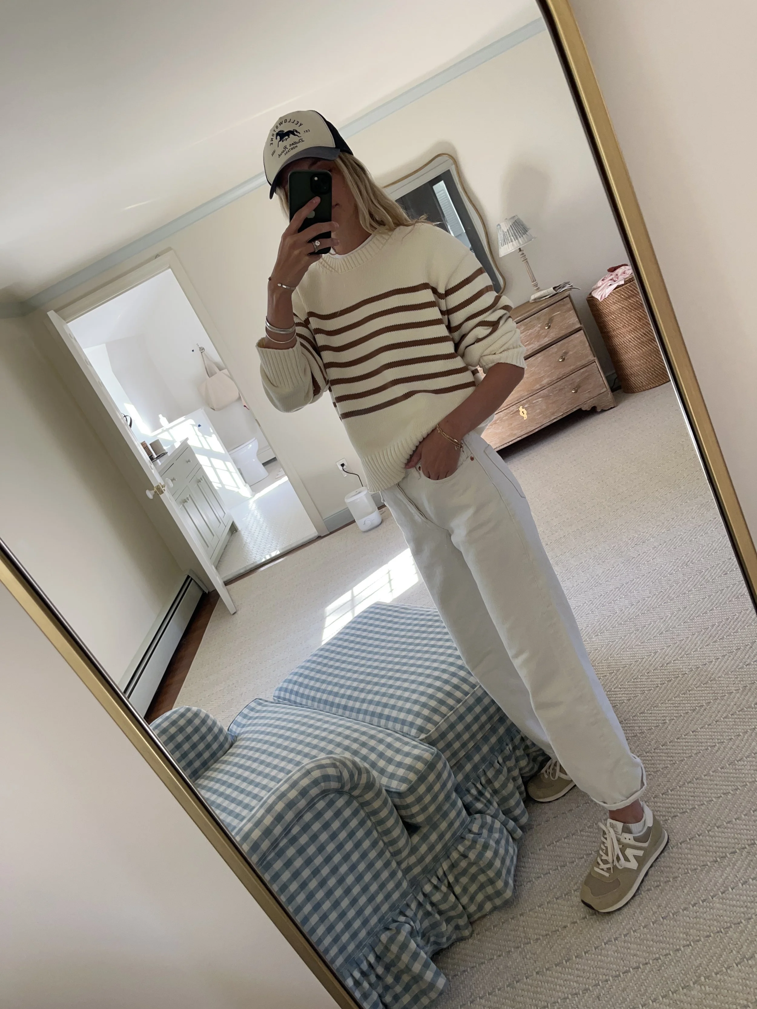 Person taking a mirror selfie in a living room with hardwood floors, wearing a black fleece jacket, gray shirt, black leggings, gray sneakers, and a white cap with a logo.