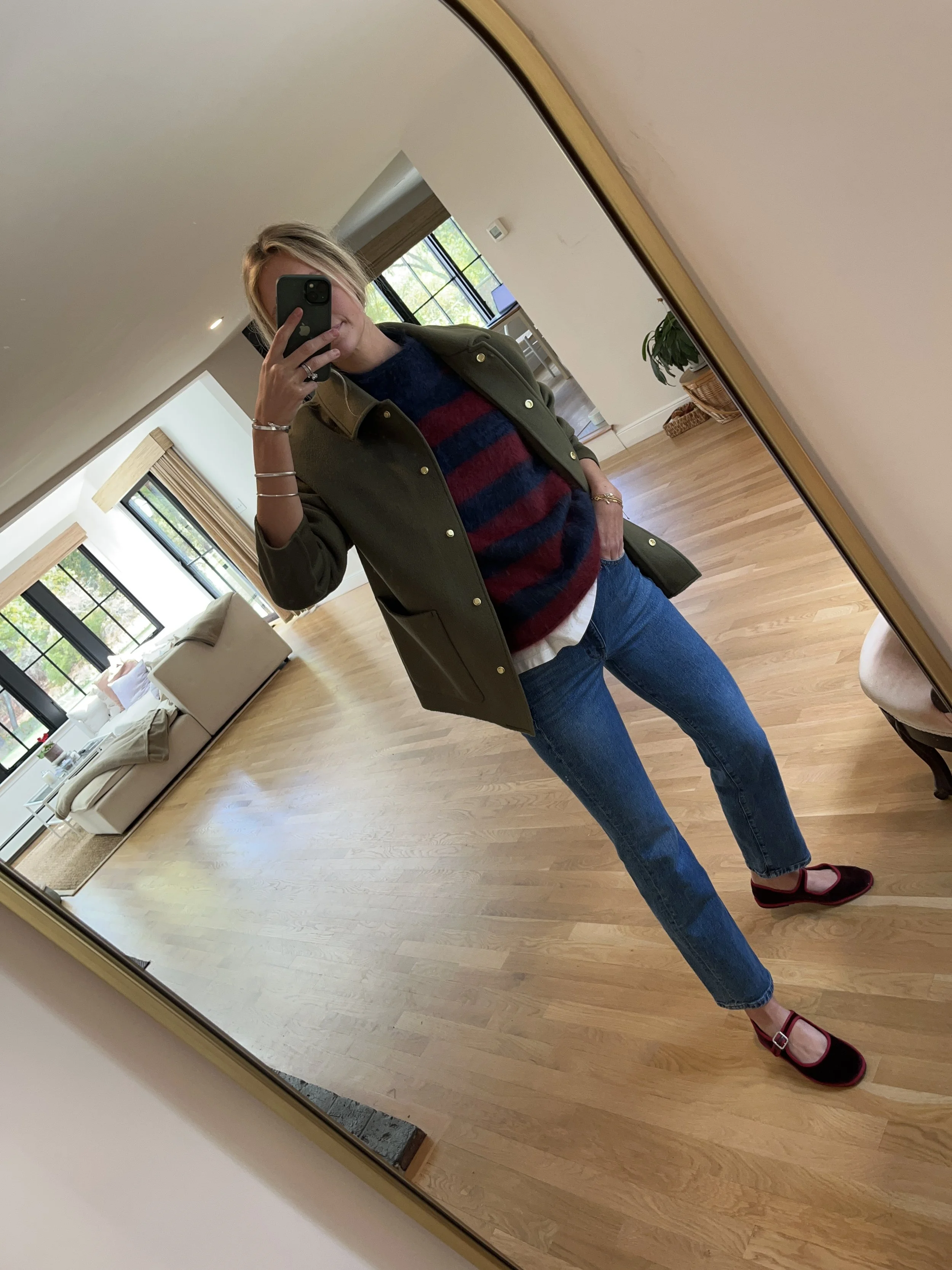 Woman taking a mirror selfie in a spacious, well-lit living room, wearing a green jacket, striped sweater, blue jeans, and burgundy shoes.