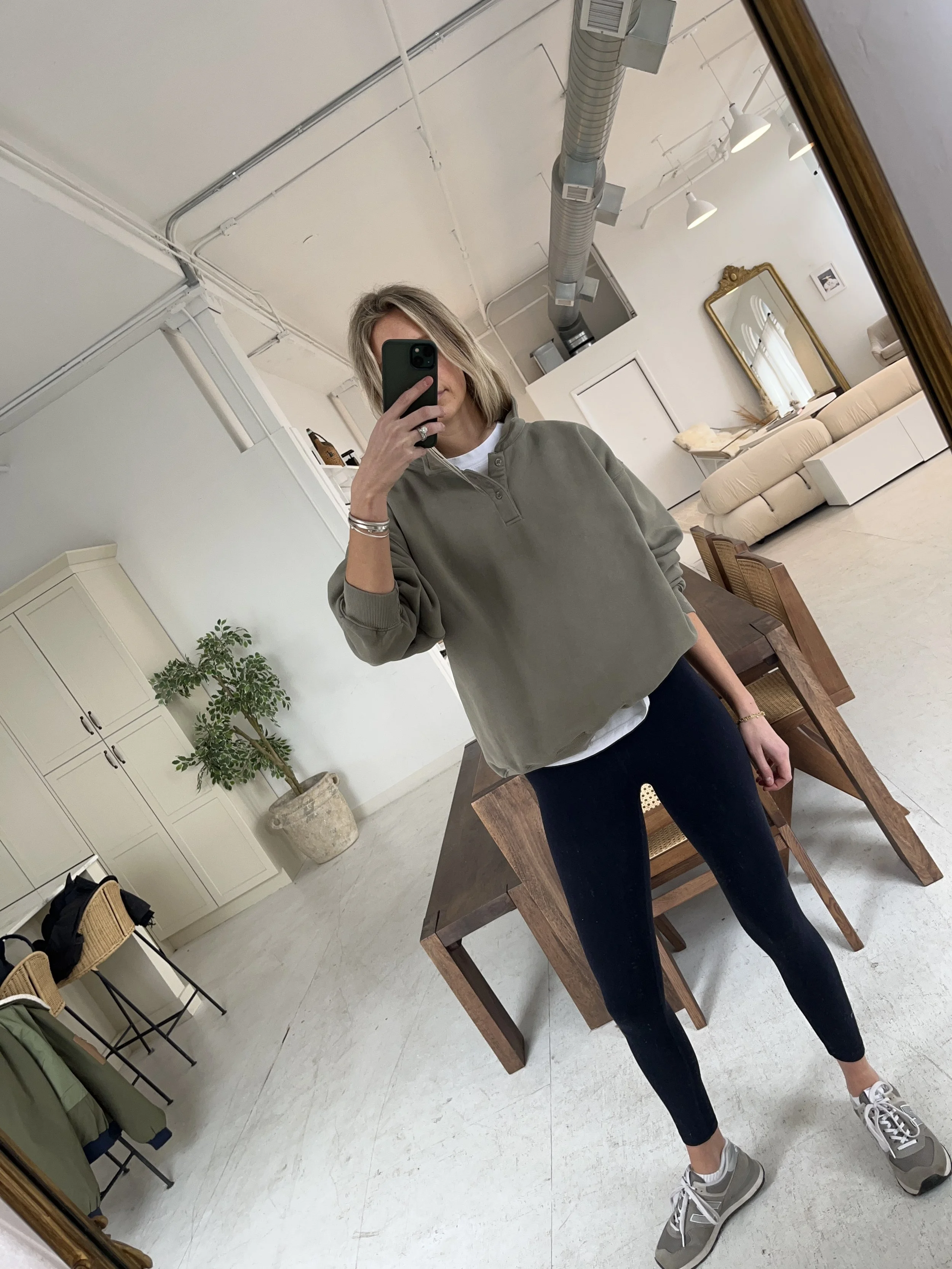 Person taking a mirror selfie in a living room with hardwood floors, wearing a black fleece jacket, gray shirt, black leggings, gray sneakers, and a white cap with a logo.