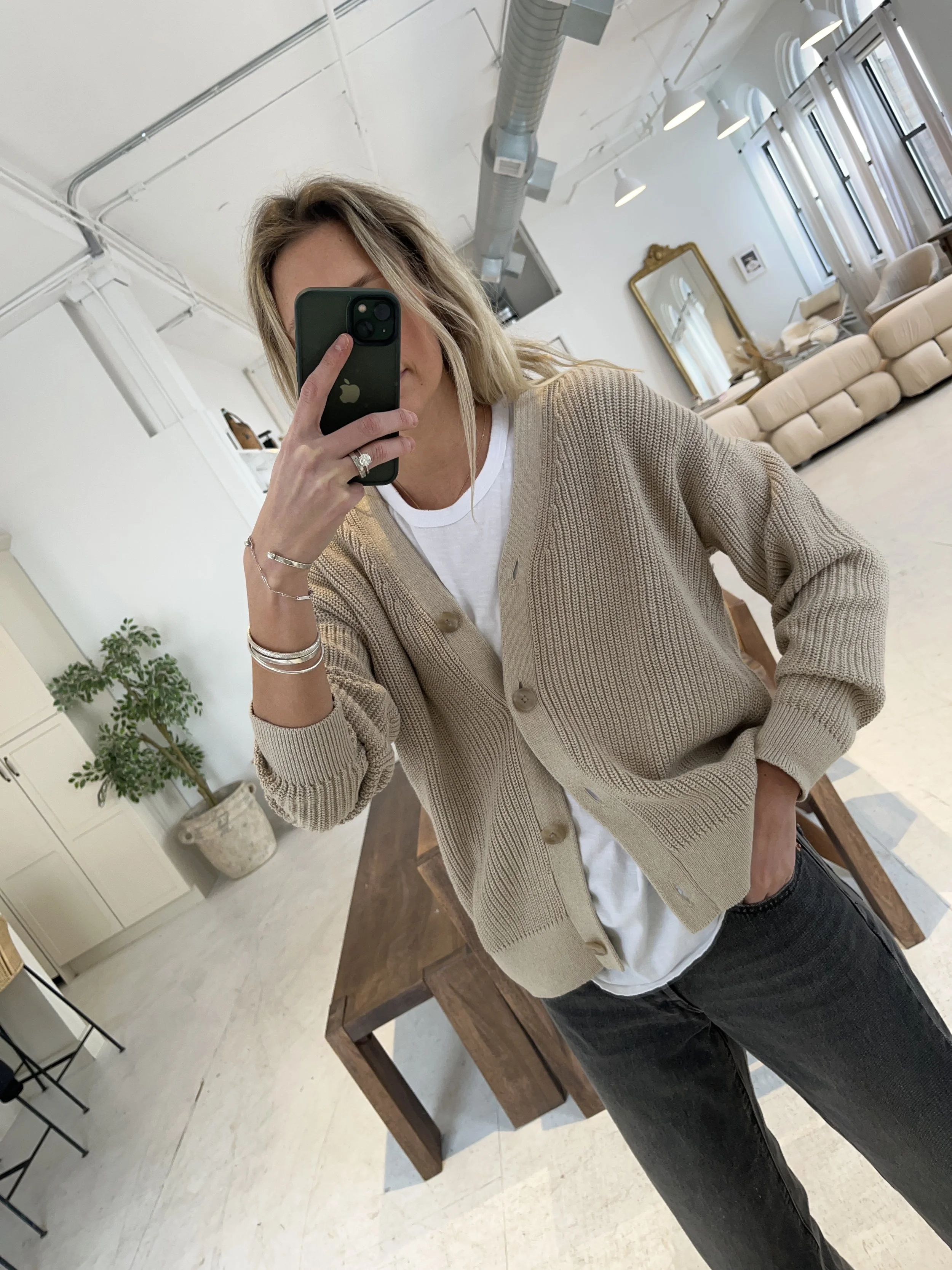 Person taking a mirror selfie in a living room with hardwood floors, wearing a black fleece jacket, gray shirt, black leggings, gray sneakers, and a white cap with a logo.