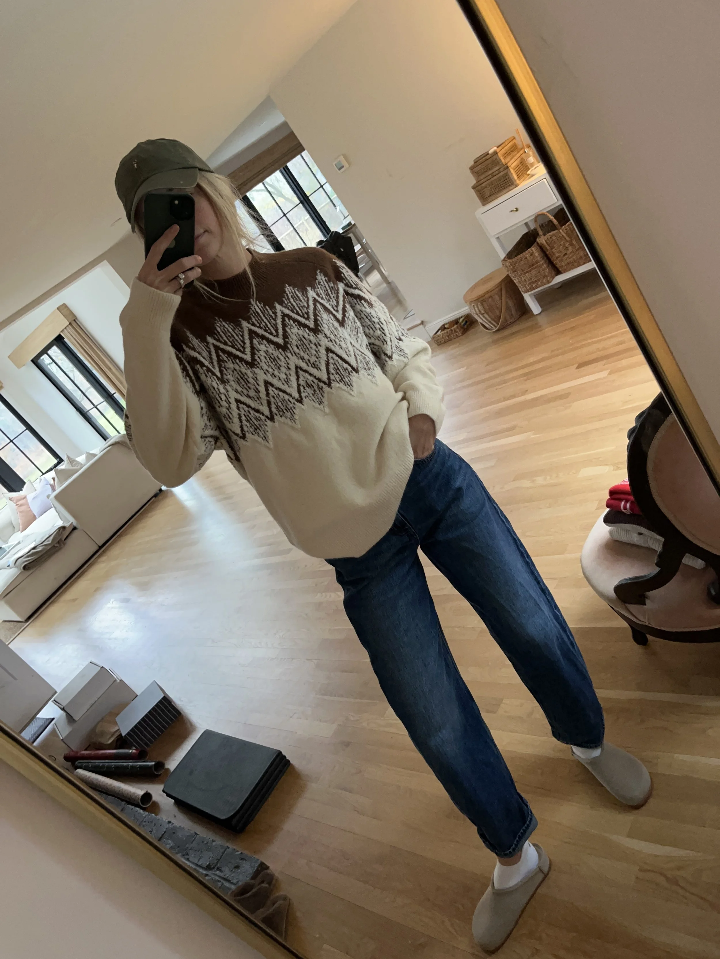 Person taking a mirror selfie in a room with wooden floors, wearing a striped brown and white long-sleeve shirt, light blue jeans, and black and white sneakers.