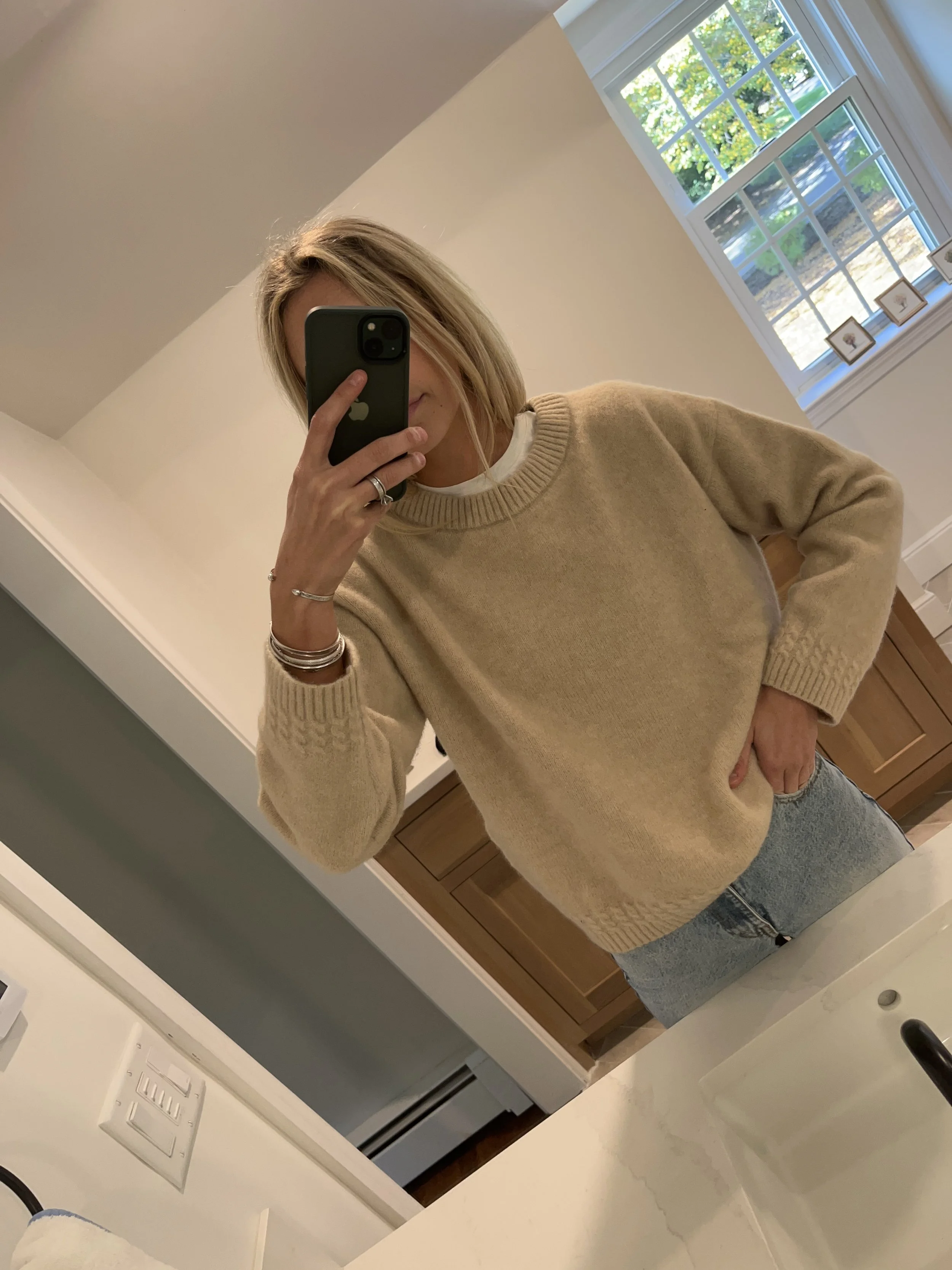 A woman taking a mirror selfie in a bathroom, wearing a beige sweater and light blue jeans, with a window showing green trees in the background.