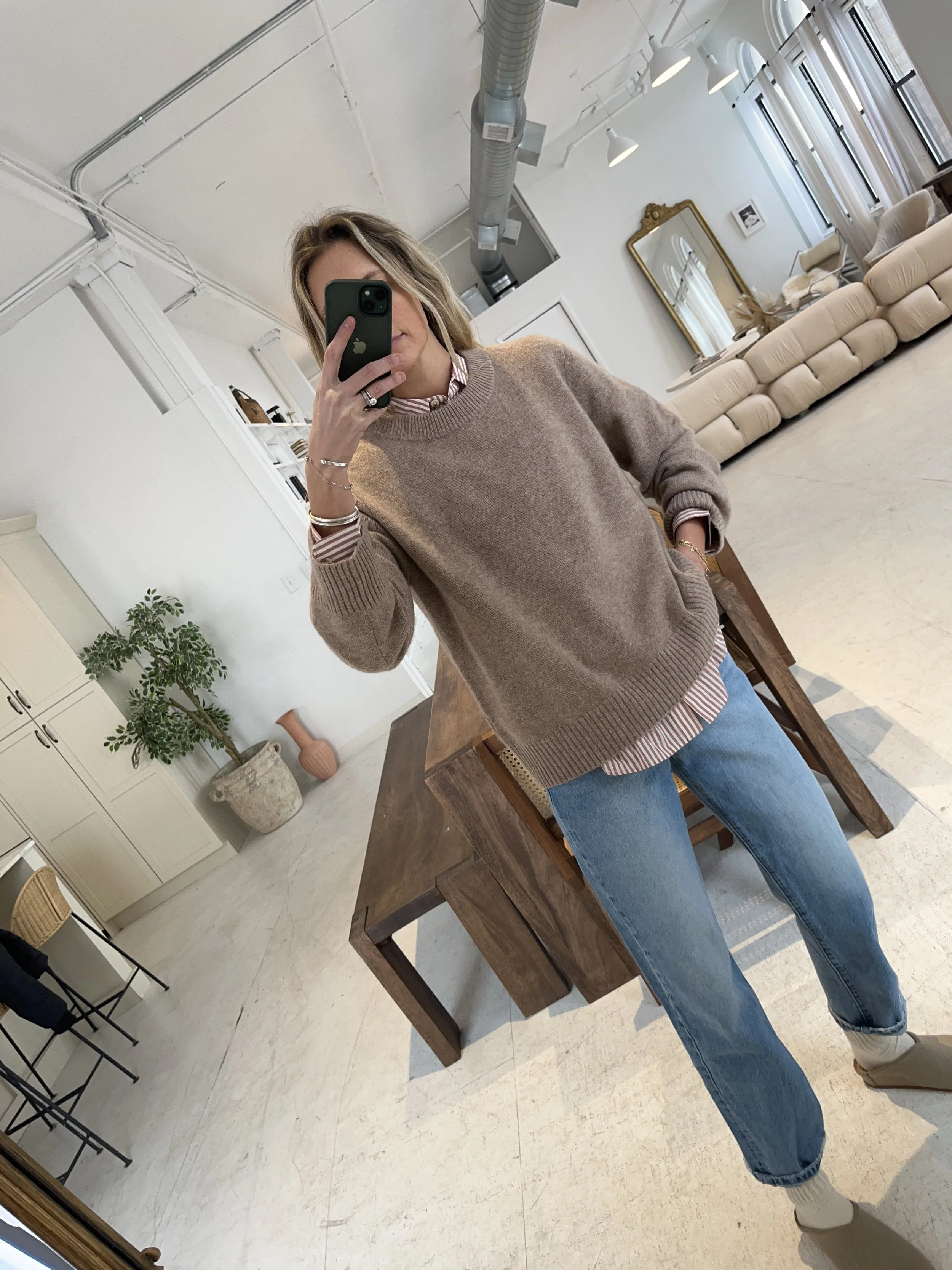 A woman taking a mirror selfie in a spacious living room with wooden floors, large windows, and minimal furniture, wearing a beige jacket, black top, blue jeans, and tan shoes.